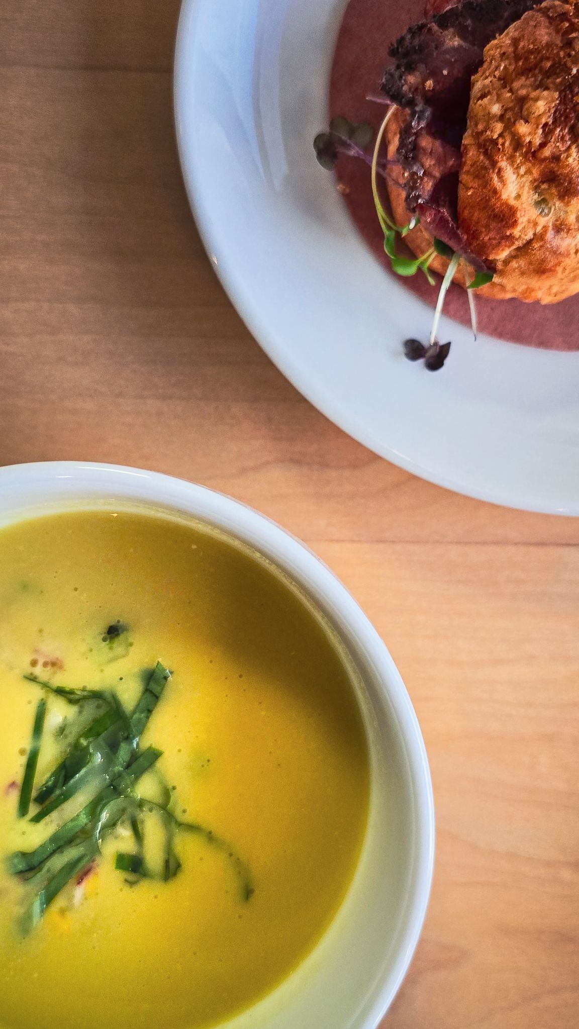 Golden yellow soup with julienned vegetables in white bowl beside a gourmet beet and fritter dish.