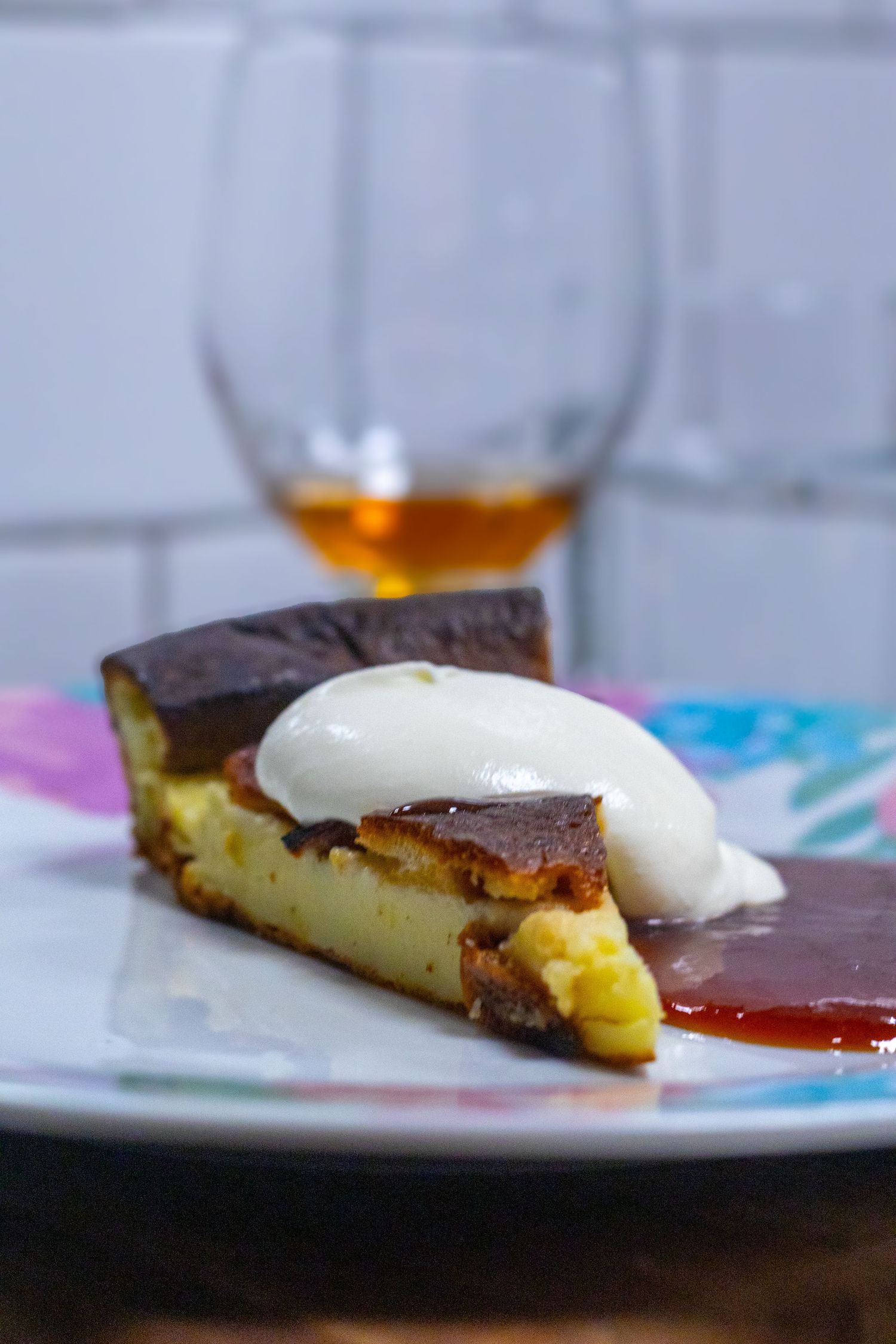 Slice of Basque burnt cheesecake topped with cream on a colorful plate, with a glass of brandy in the background.