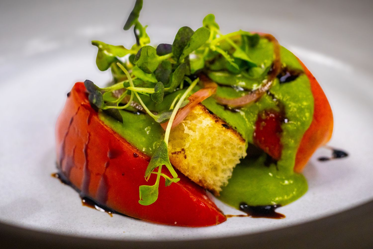 Gourmet tomato salad with green pea puree, microgreens, and balsamic glaze on a white plate.