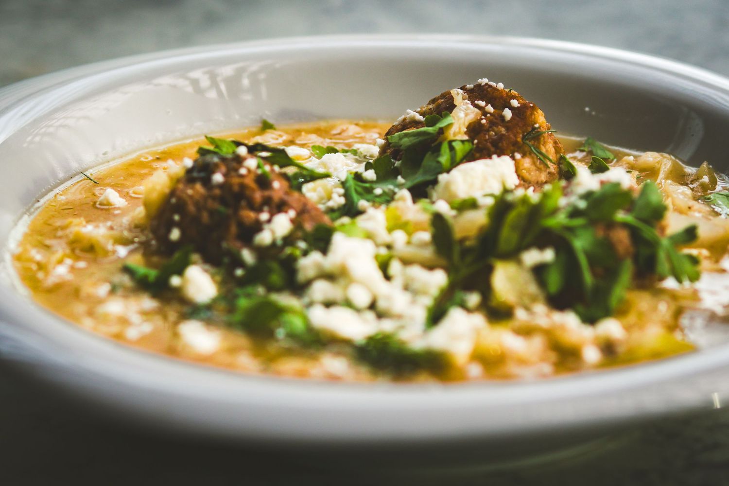 A white bowl of creamy soup topped with crumbled feta, fresh herbs, and braised meat garnish.
