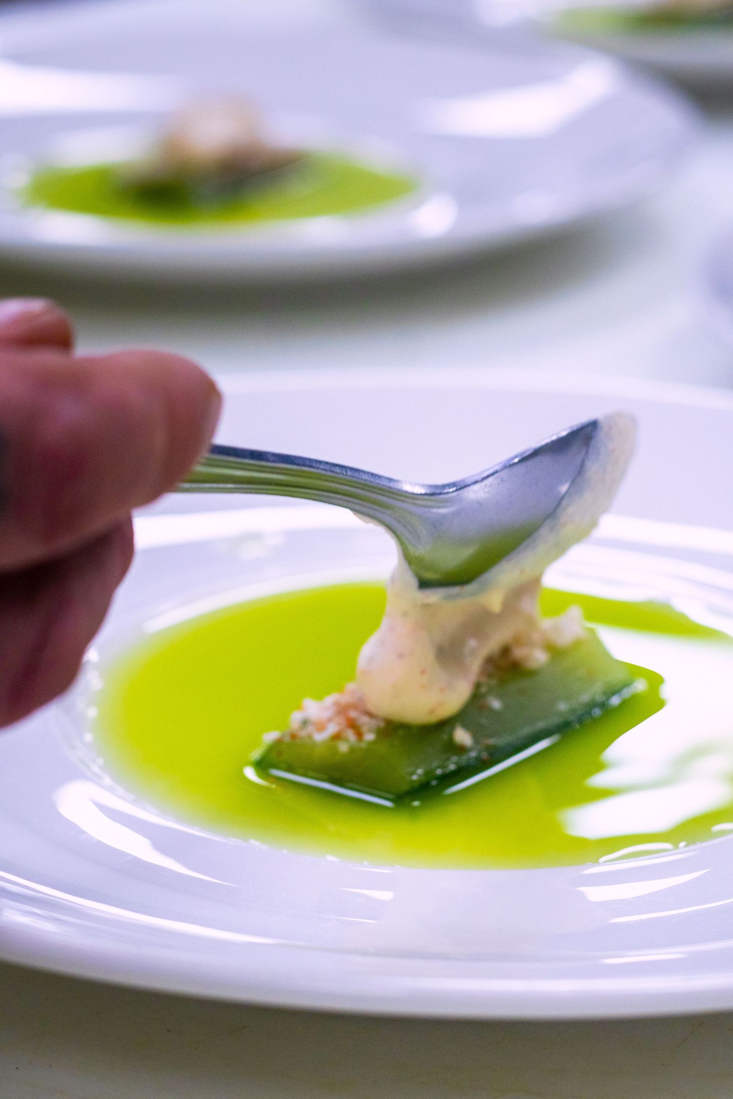 Chef plating a gourmet dish with creamy sauce on green pasta in vibrant herb oil on a white plate.