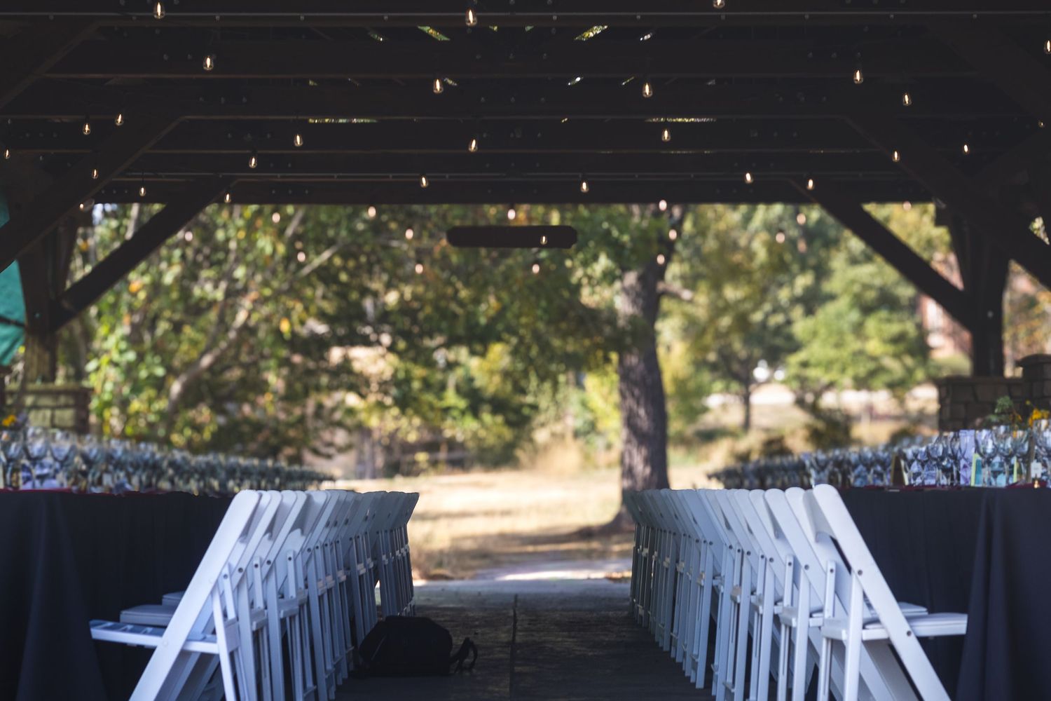 An elegant outdoor wedding venue setup with white chairs lining an aisle under a covered pavilion with string lights.