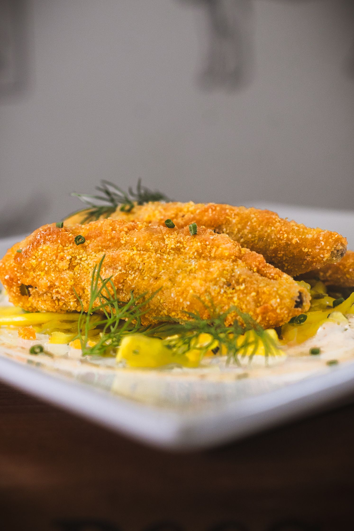 Golden breaded fish fillets served on creamy mashed potatoes, garnished with fresh herbs and yellow sauce on a white plate.