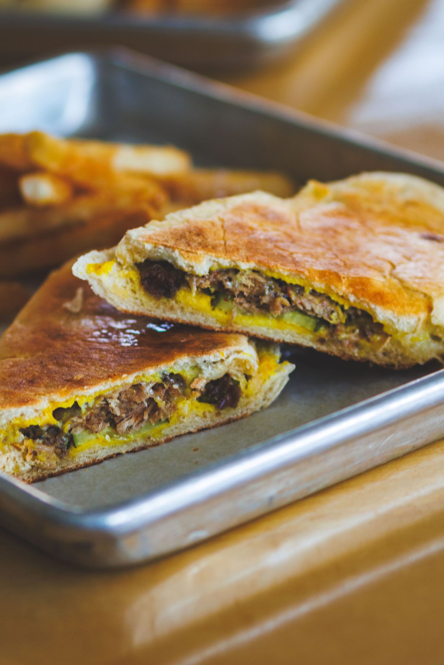 Toasted flatbread sandwich filled with seasoned beef and melted cheese, served halved on a metal tray with fries.