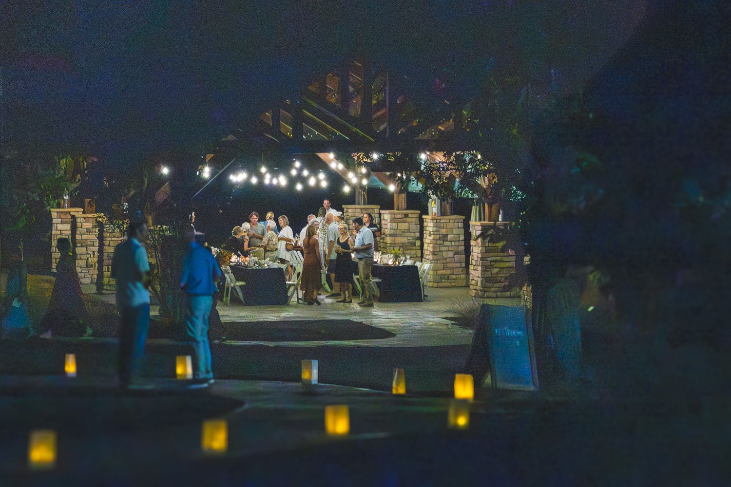 Outdoor evening gathering with string lights and candle luminaries lining a patio at a rustic venue.