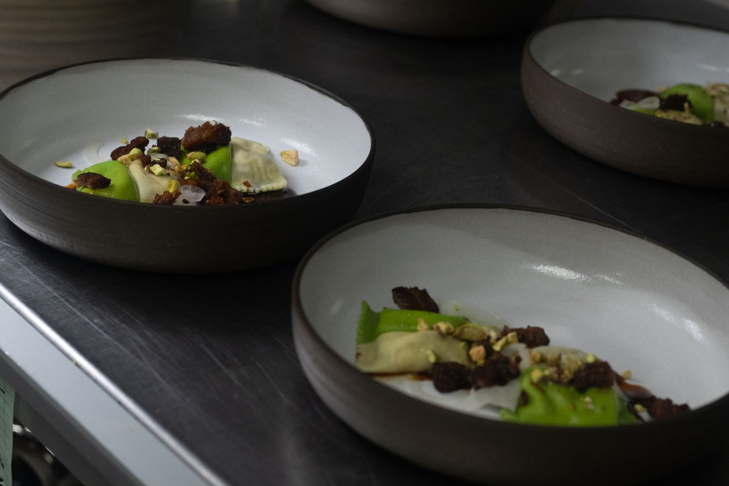 Three modern gray bowls plated with gourmet ingredients including greens, chocolate crumbles, and cream on a dark surface.