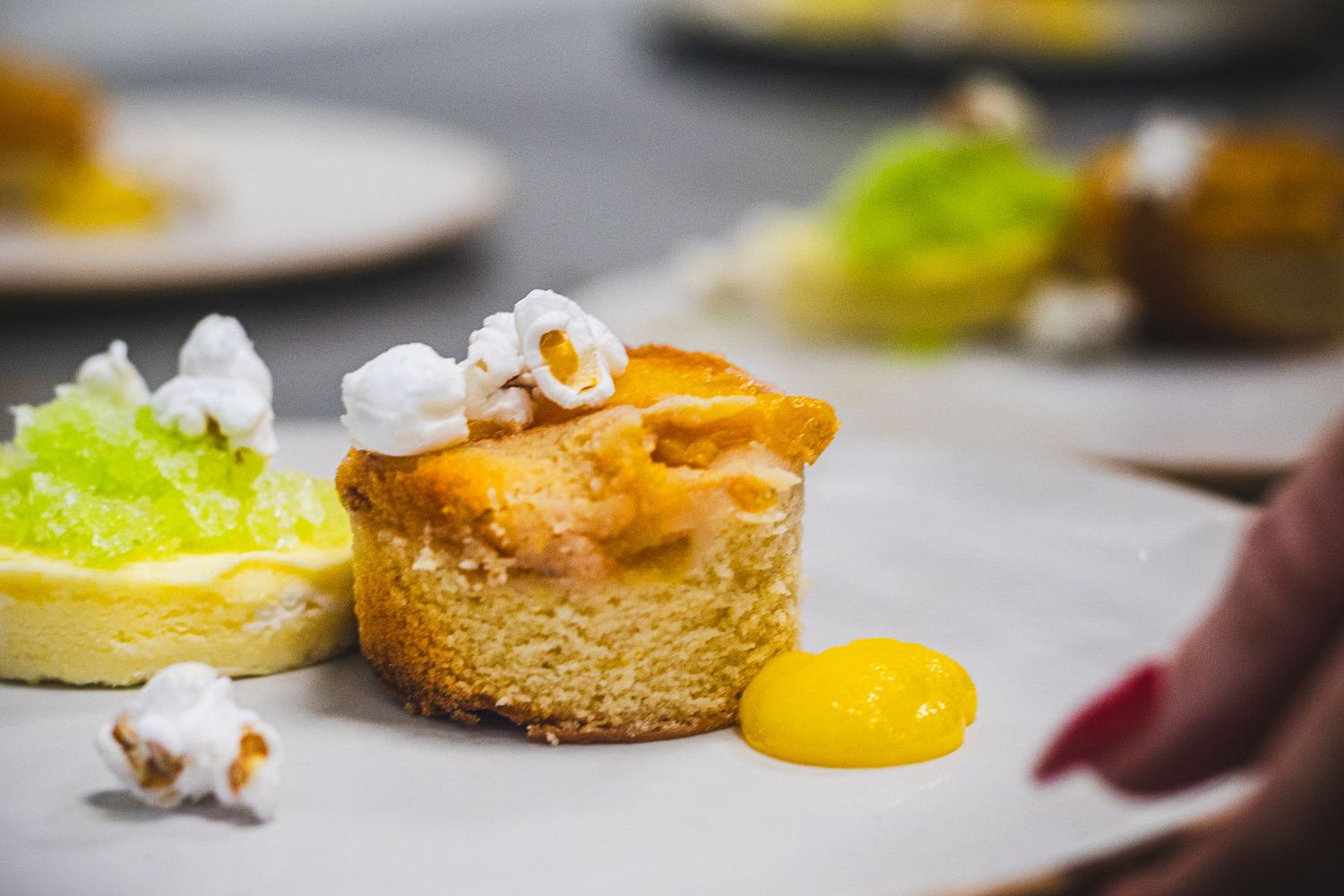 Elegant plated dessert with a golden mini cake, mango coulis, popcorn garnish, and green sorbet on a white plate.