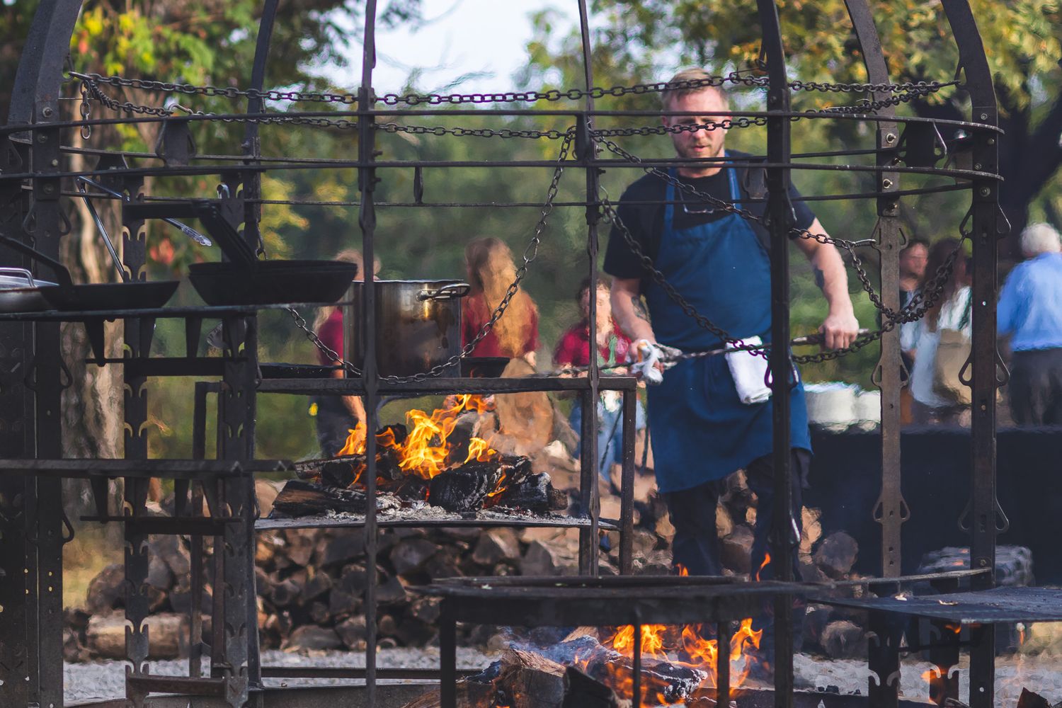Chef cooking over open fire on large outdoor grill at a rustic culinary event.