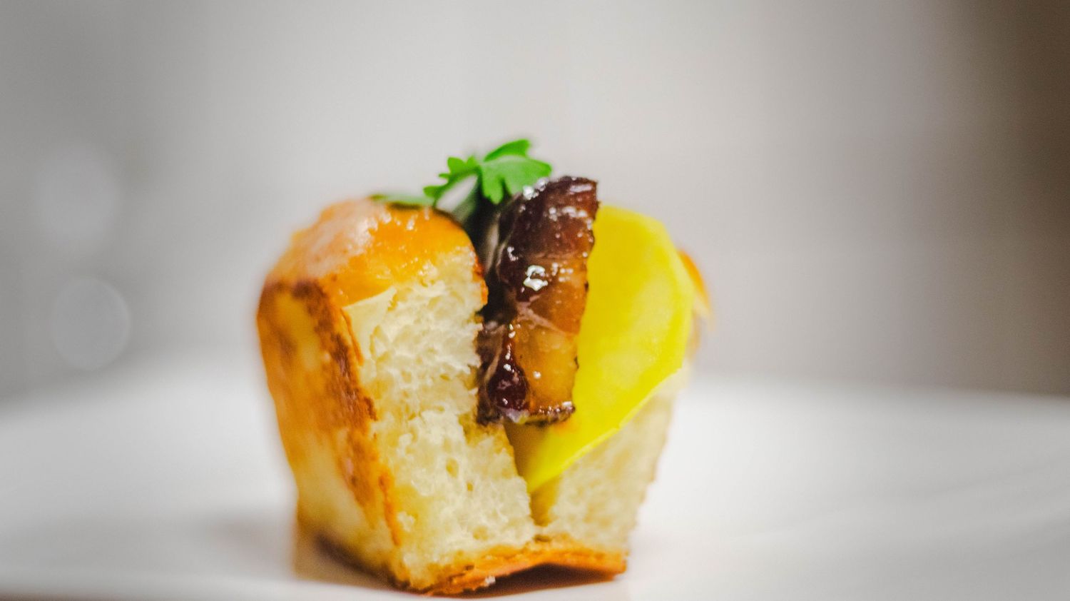 Gourmet brioche bun topped with glazed meat, yellow sauce, and fresh herbs on a white plate.