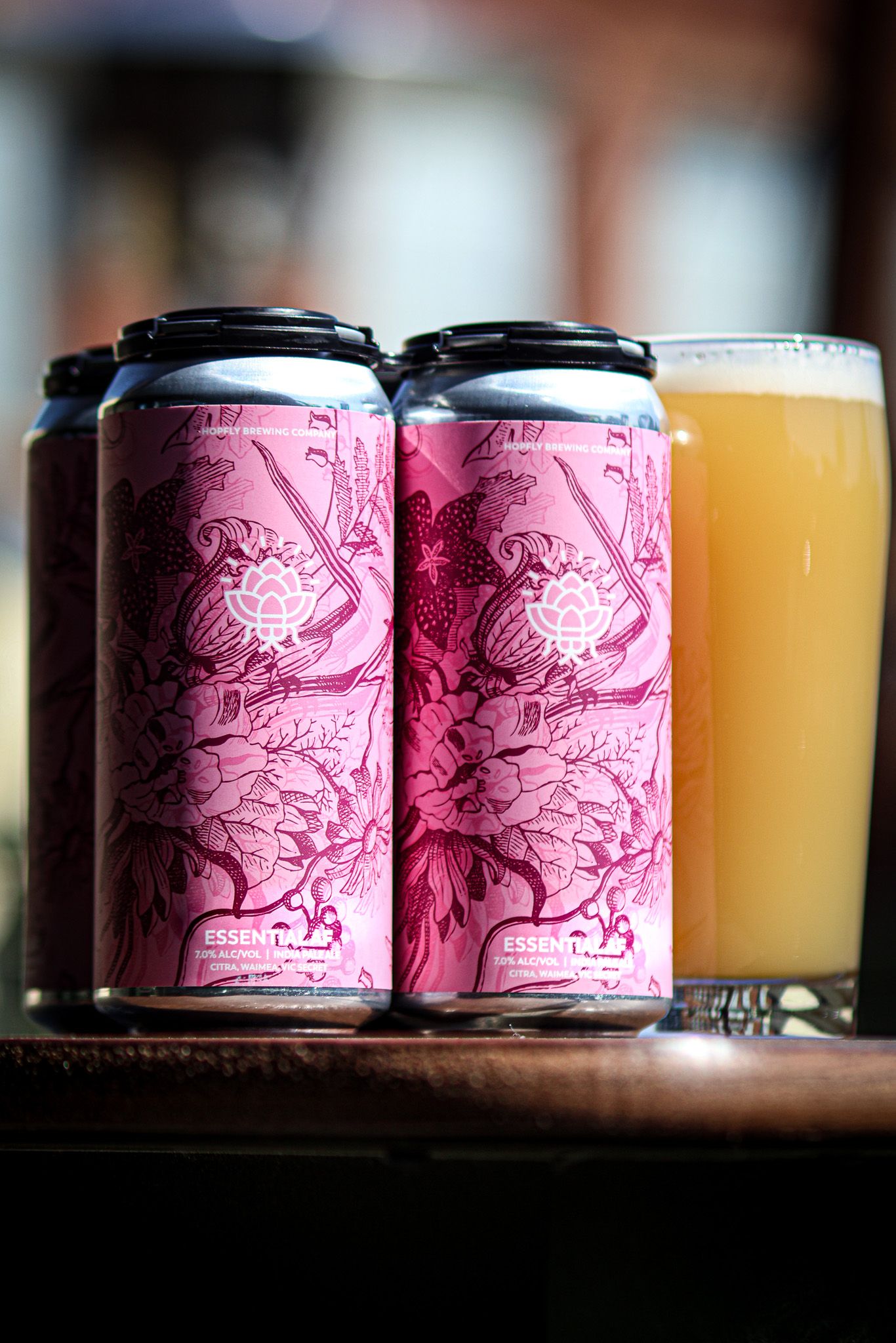 Four pink cans of Essential craft beer beside a glass of hazy IPA on a wooden surface.