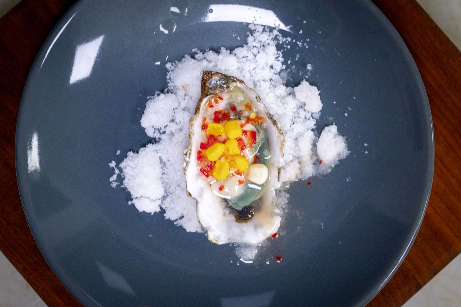 Fresh oyster on the half shell served on a bed of rock salt with colorful pepper garnish on a blue plate.