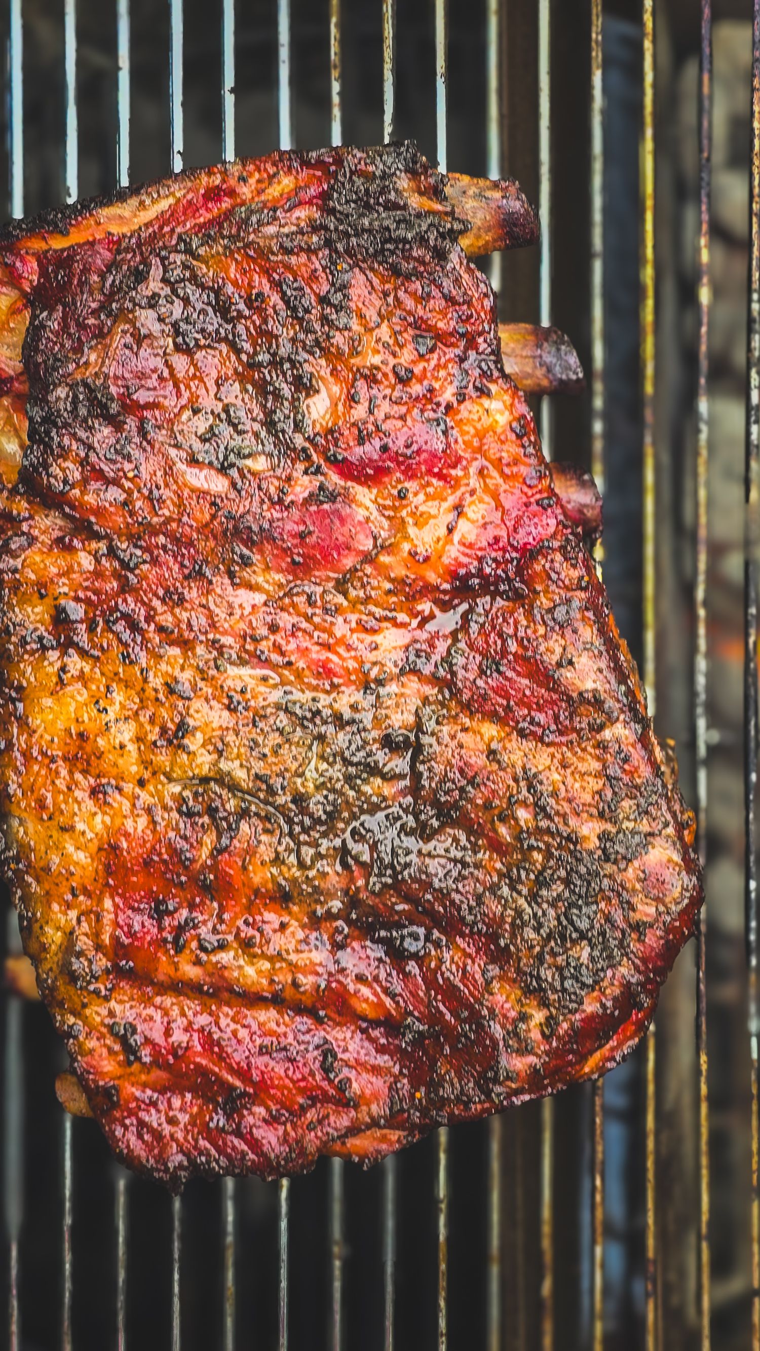 Close-up of a smoky BBQ pork shoulder with charred crust cooking on a grill, showcasing rich red and brown tones.
