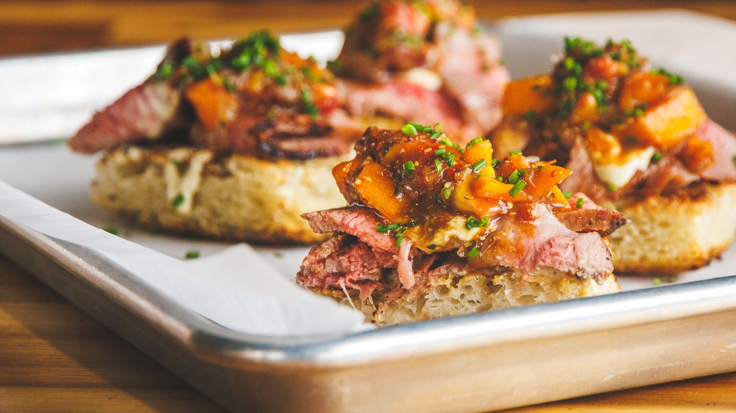 Toasted bread topped with tender pulled meat, roasted vegetables, and fresh herbs on a metal tray.