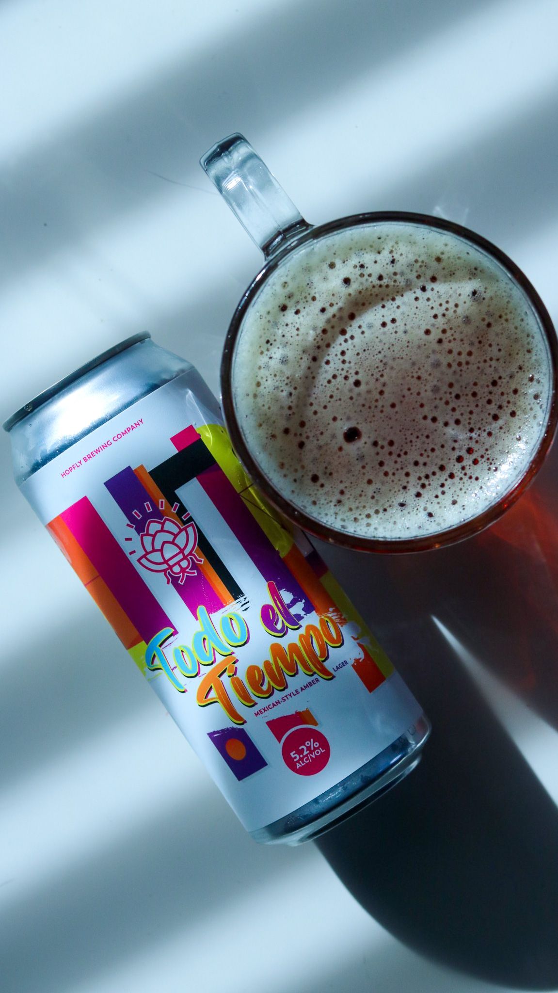 Colorful Todo el Tiempo beer can beside a frothy pint glass on a white surface with dramatic shadows.