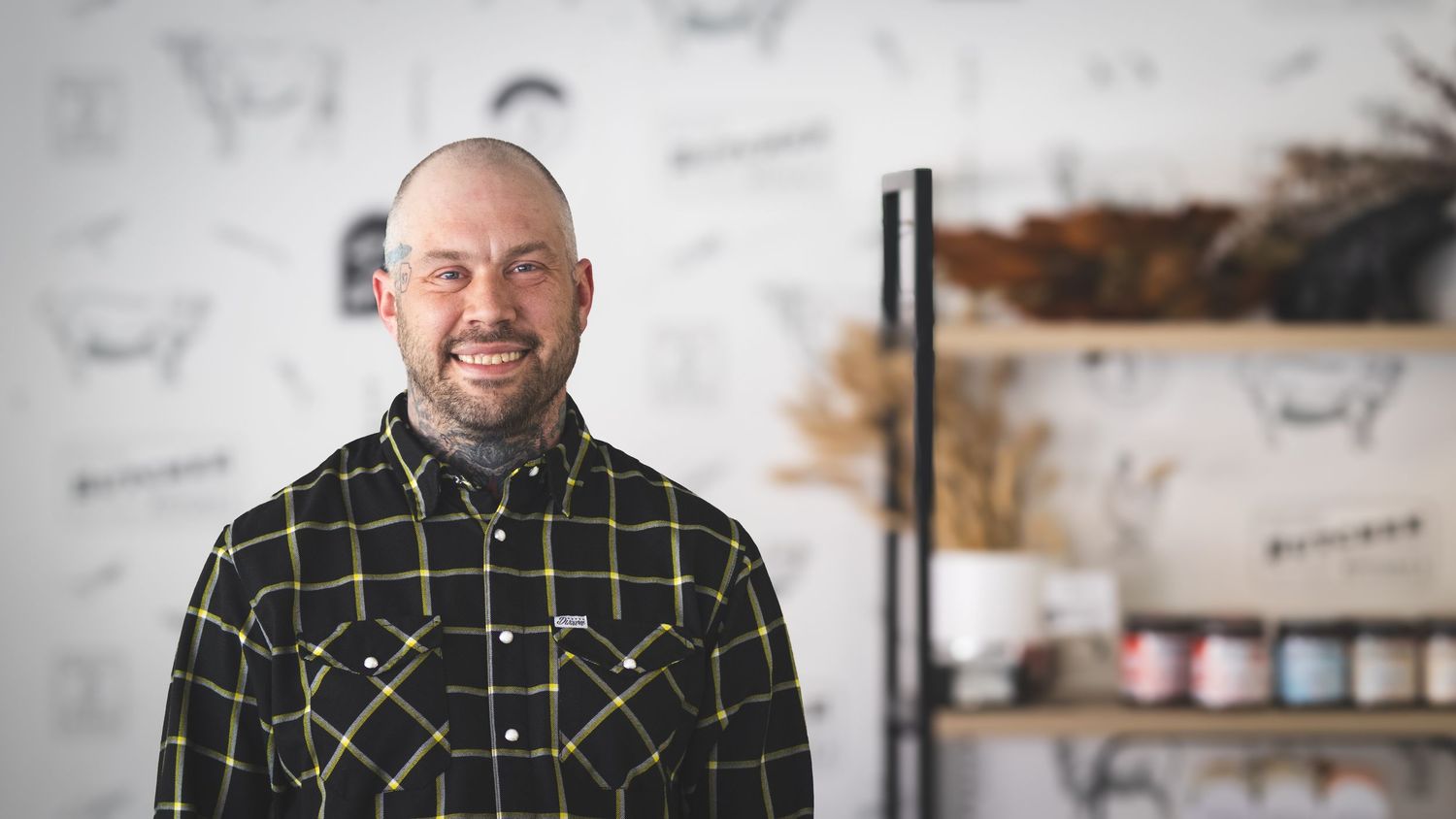Smiling bald man with beard wearing plaid shirt stands in modern barbershop with branded decor.