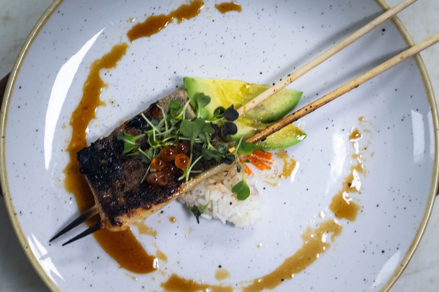 Glazed fish fillet served over rice with avocado, microgreens, and savory brown sauce on white plate.