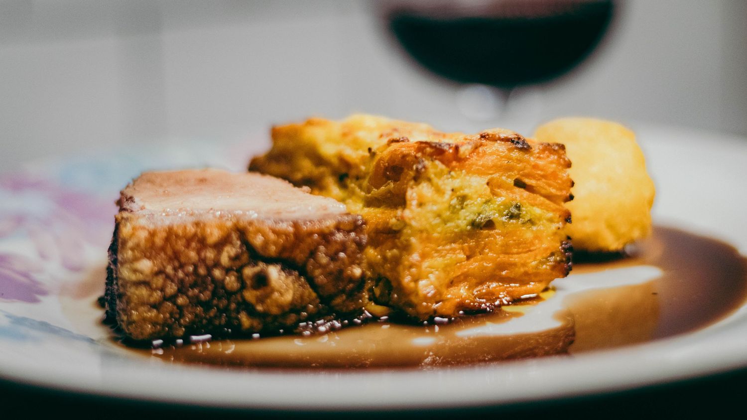 Seared meat slice with crispy vegetable cake served in rich brown sauce on a decorative plate with wine.