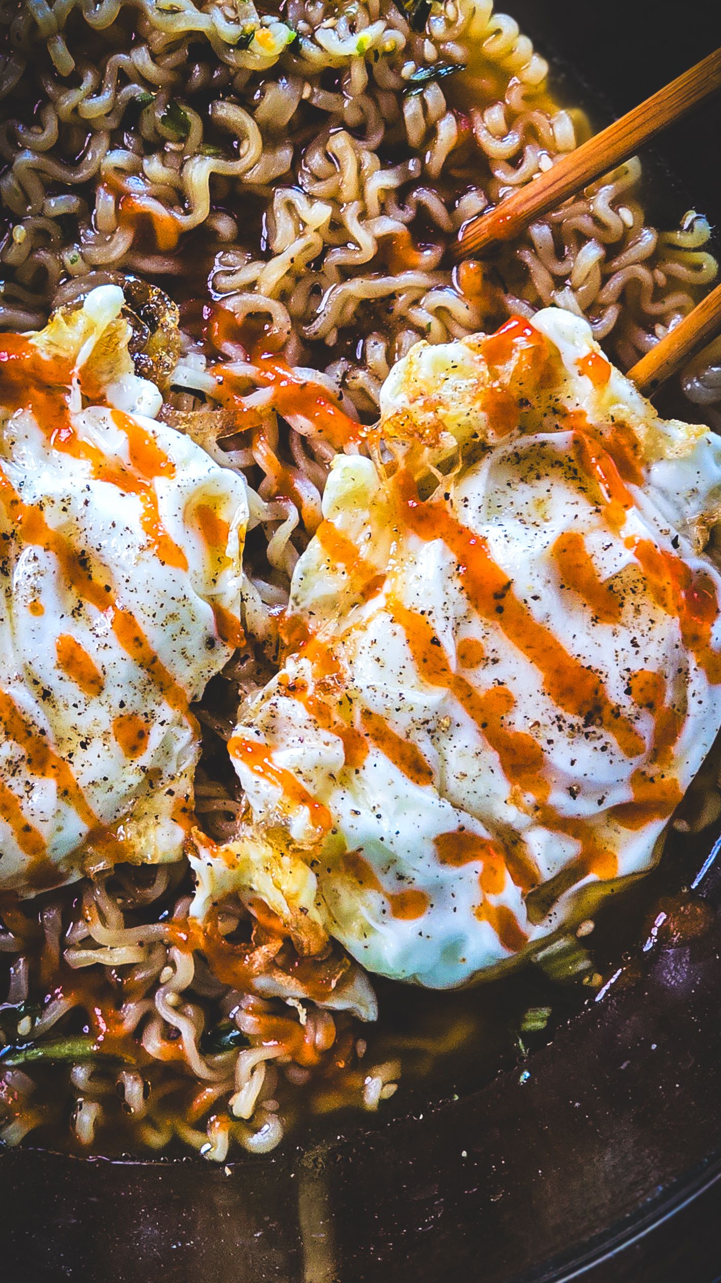 Spicy ramen noodles topped with fried eggs drizzled with sriracha sauce and black pepper in a dark skillet.