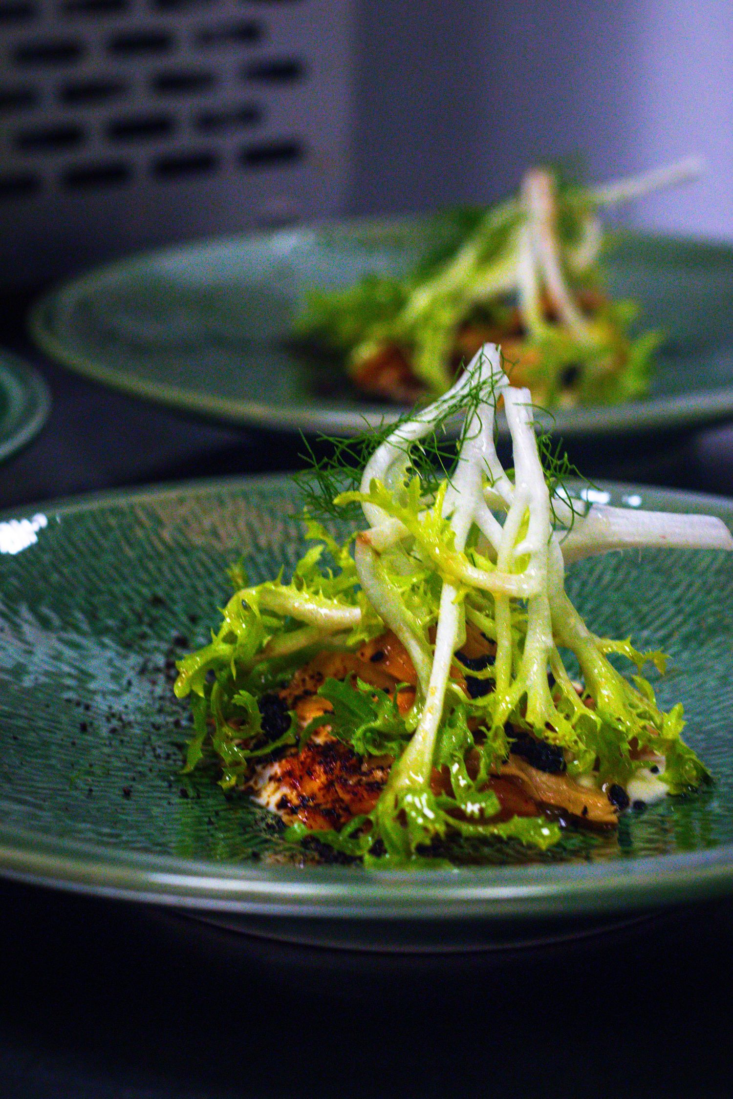 Elegant green ceramic plate with fresh frisée salad, fennel, and seasoned fish at a fine dining restaurant.