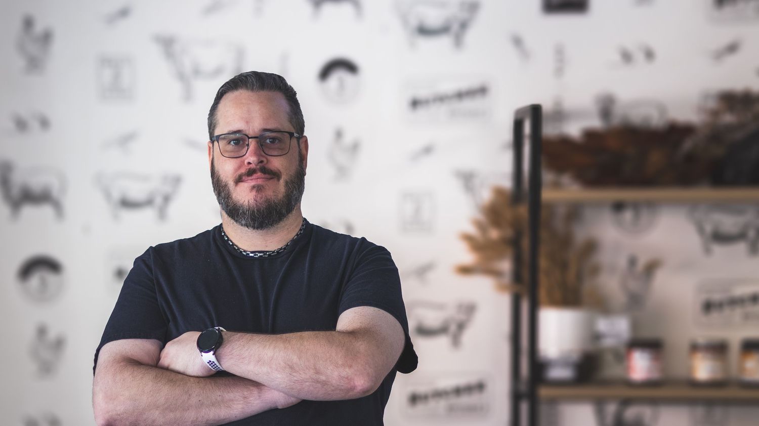 A smiling bearded man wearing glasses and a black shirt stands with arms crossed in a modern restaurant.