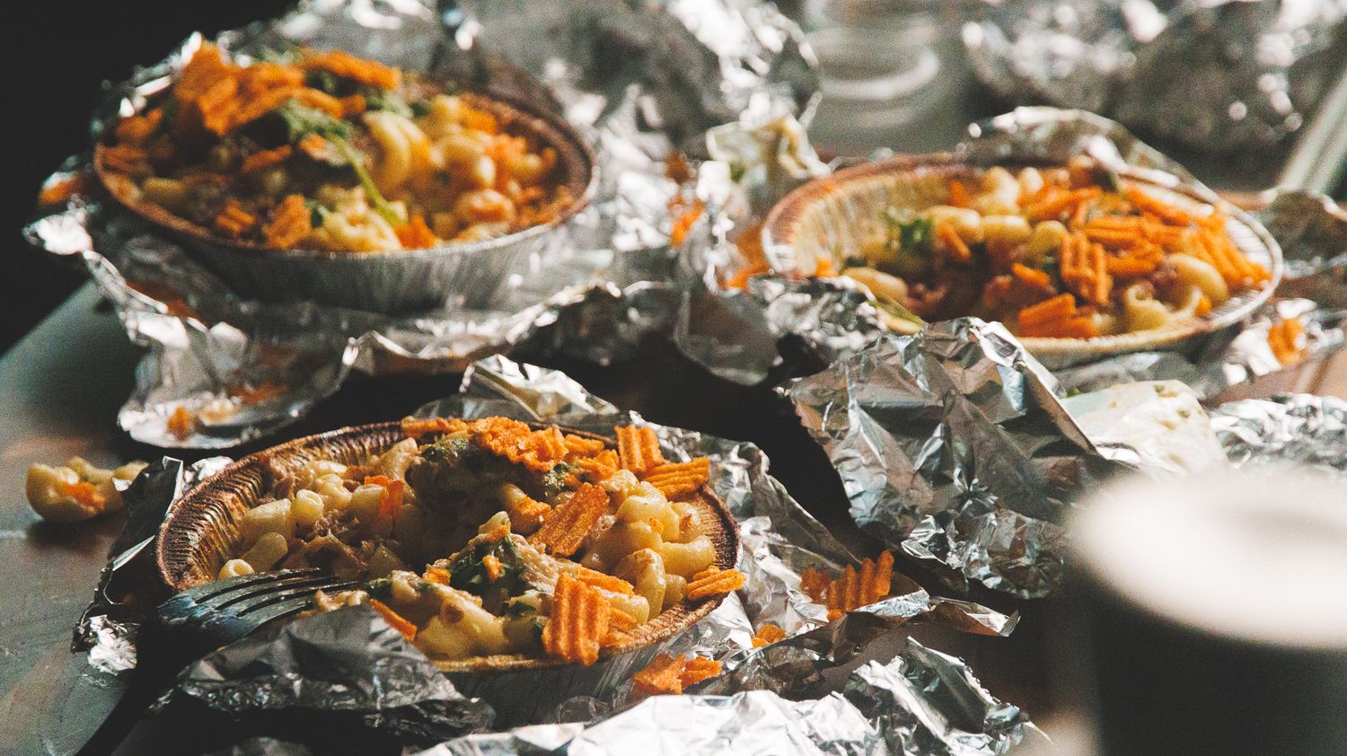 Foil-wrapped dishes filled with colorful roasted vegetables and waffle fries served as individual portions on a table.