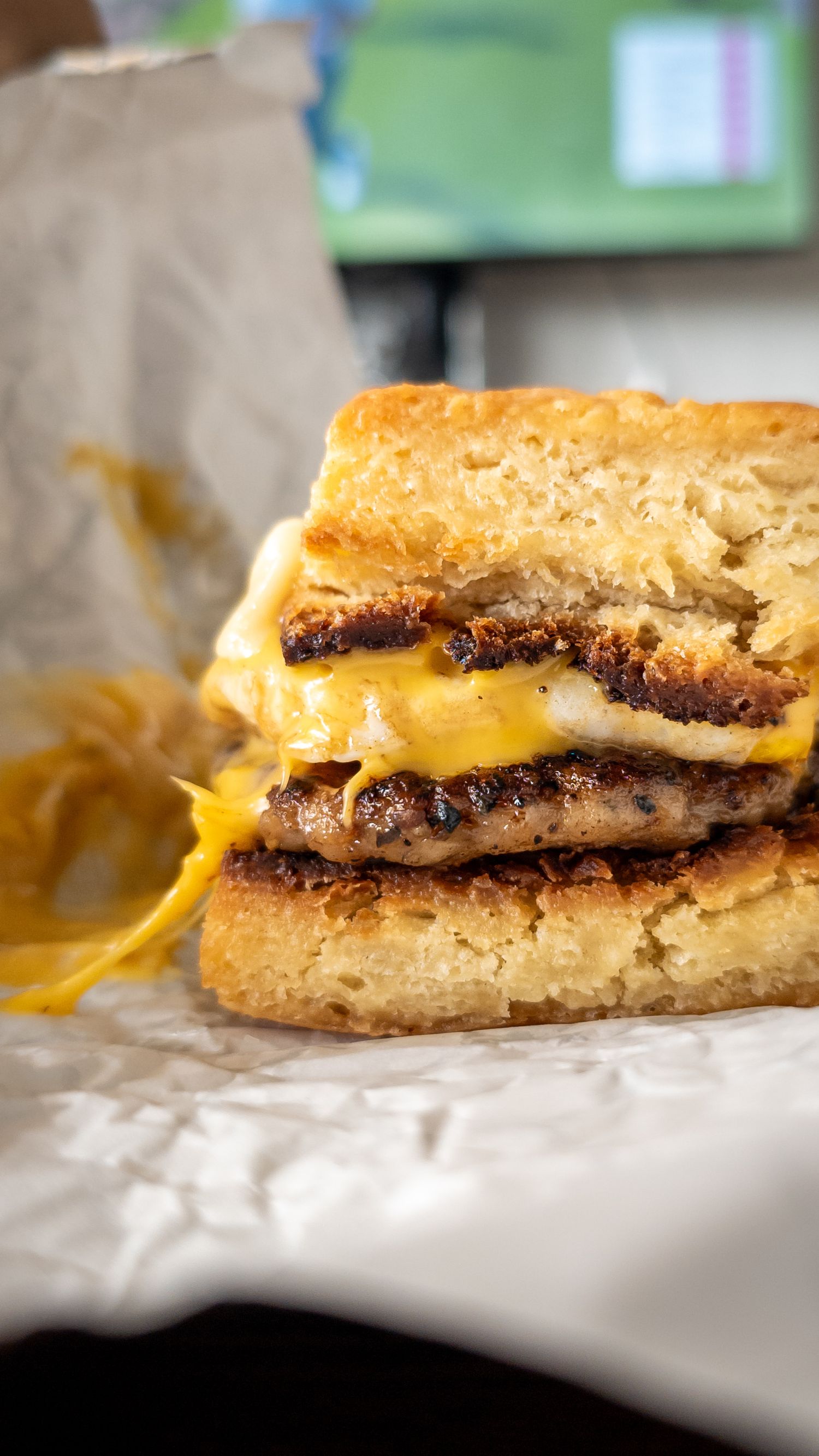 A golden biscuit breakfast sandwich with melted cheese and sausage patty on wax paper next to a paper bag.