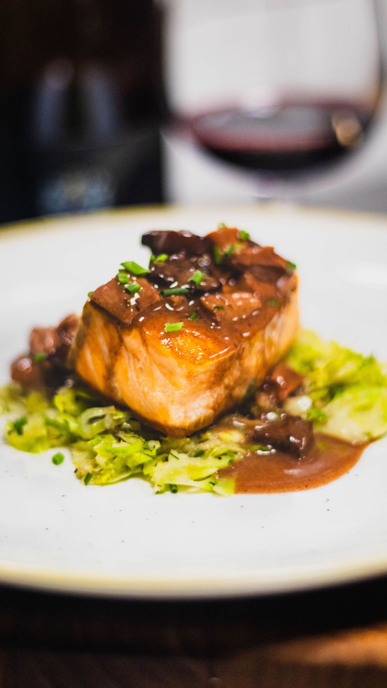 Pan-seared salmon fillet topped with mushroom red wine sauce on shredded Brussels sprouts with a wine glass background.