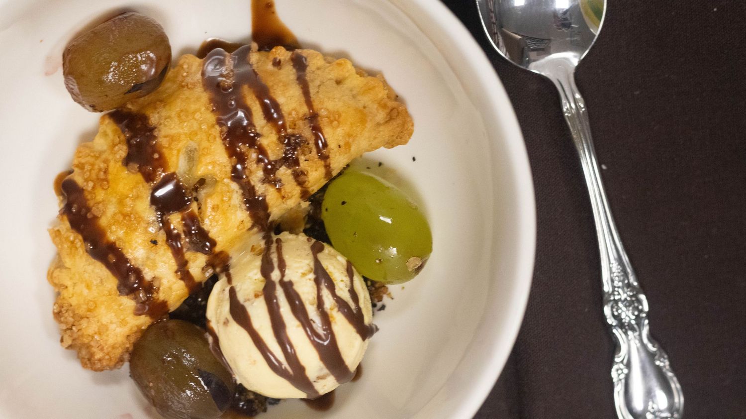 Golden fried dessert topped with chocolate drizzle, vanilla ice cream, and fresh grapes served on a white plate.