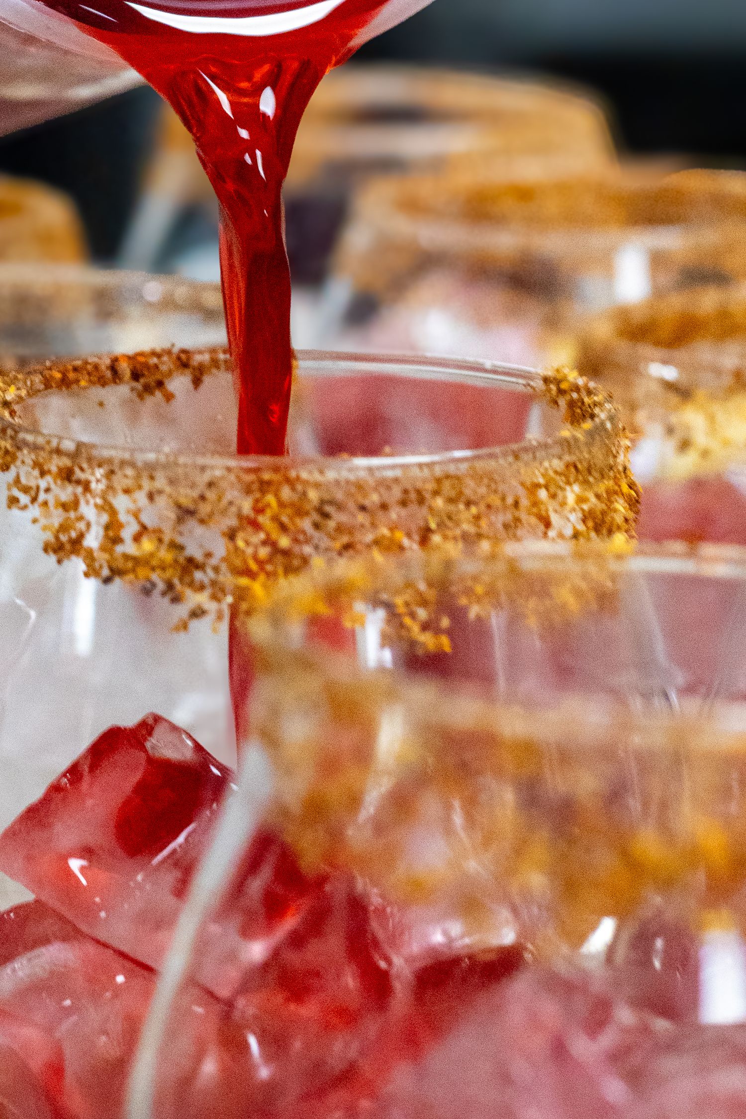 Red cocktail being poured into glasses rimmed with golden sugar crystals and filled with ice cubes.
