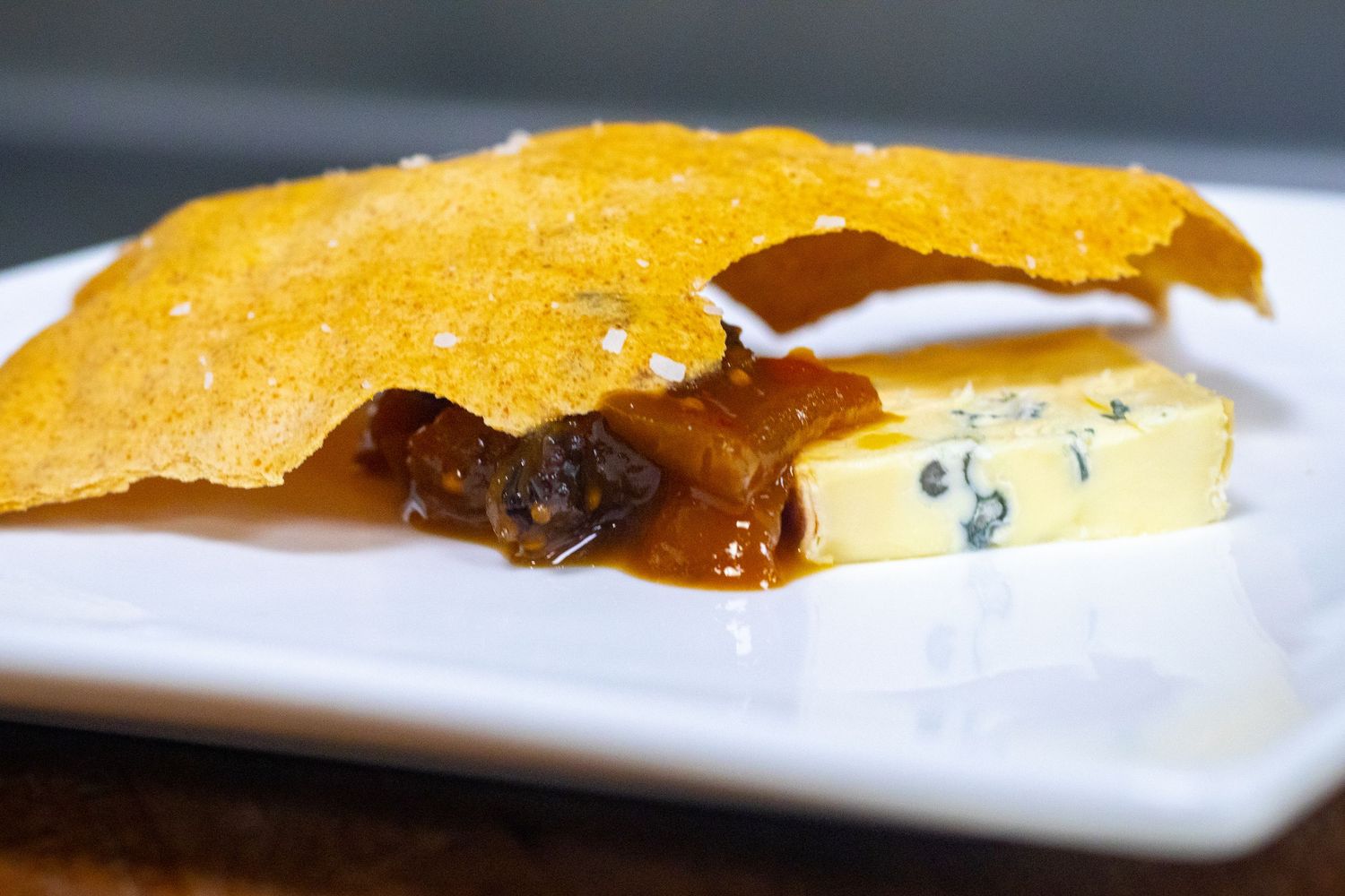 Elegant cheese plate with blue cheese, caramelized onion jam, and golden crispy cracker on white dish.