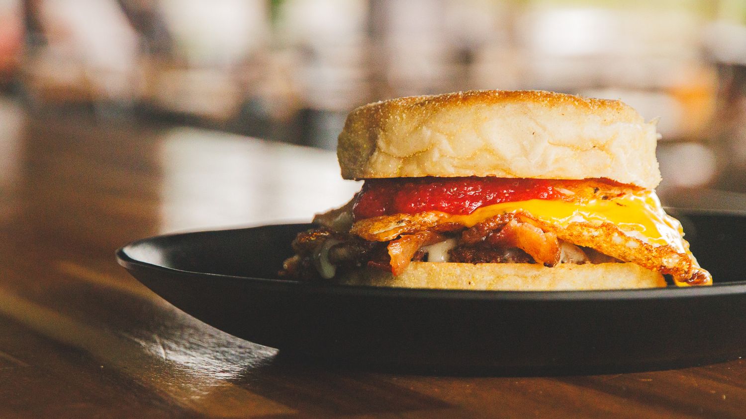 A breakfast sandwich with bacon, egg, and cheese on a toasted English muffin served on a black plate.