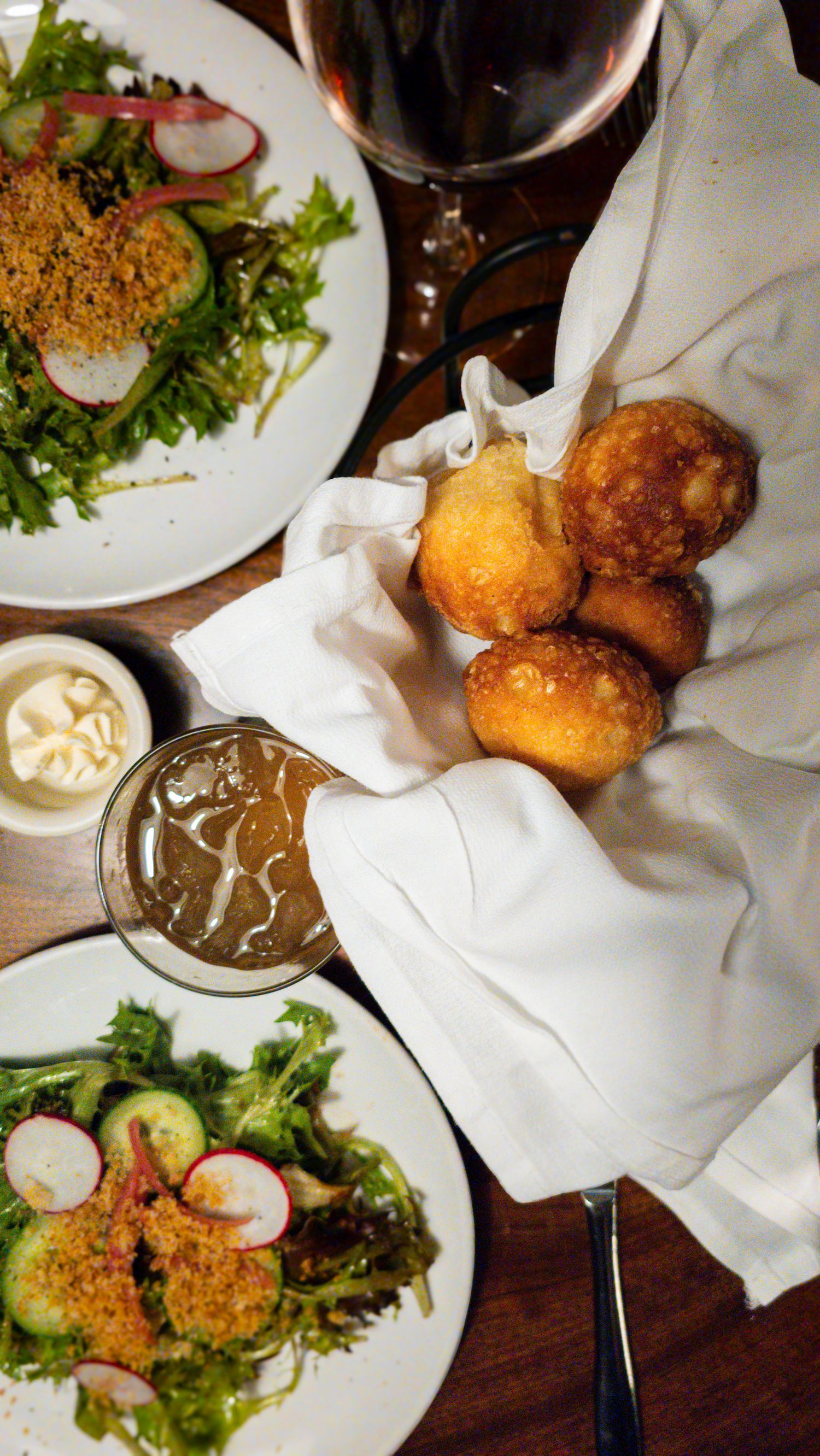 Restaurant table with fried dough balls in white cloth, green salads with radishes, dipping sauce, and red wine.