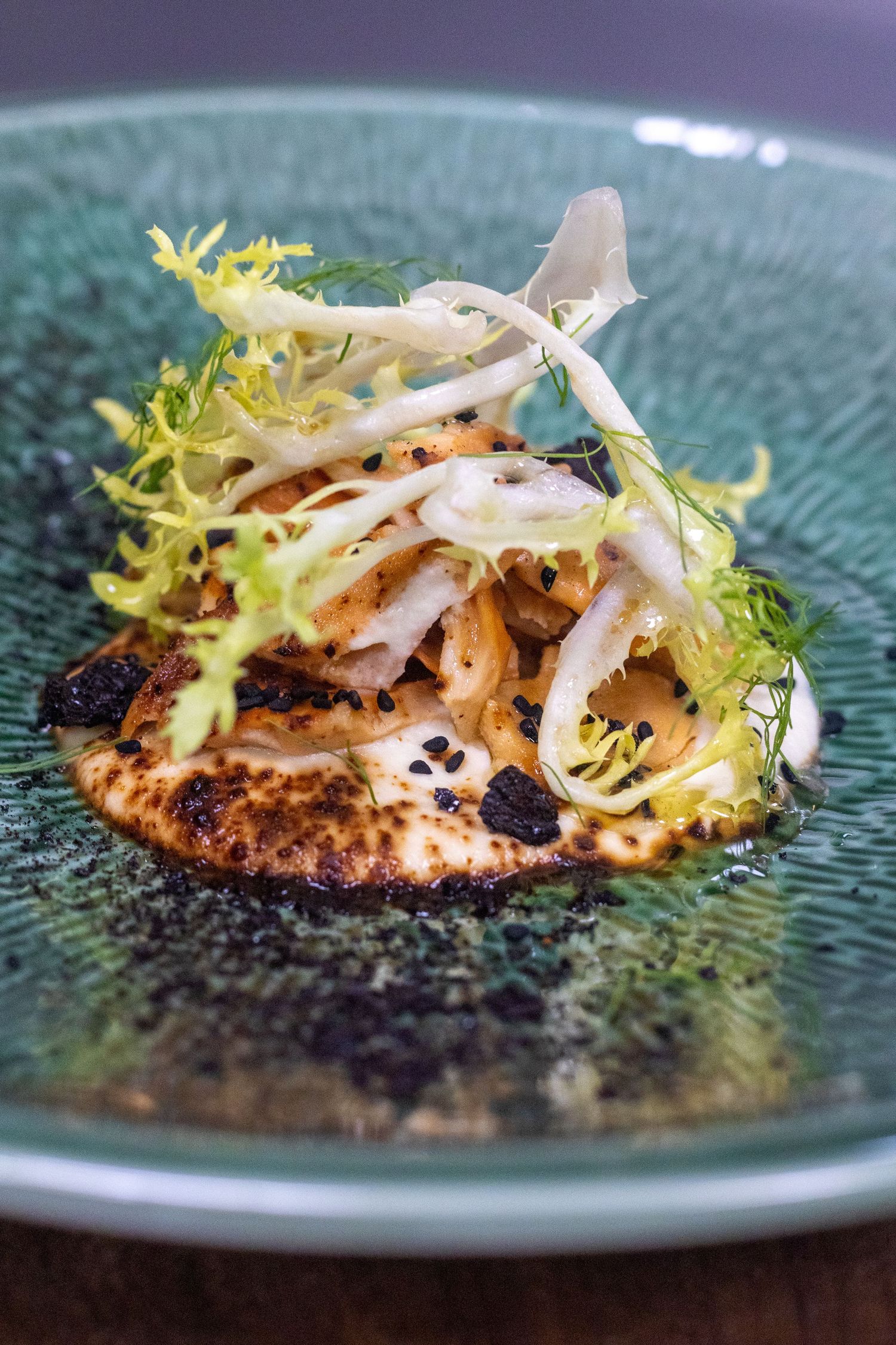 Elegant fine dining dish with sautéed seafood, frisée lettuce, and black sesame on a textured teal ceramic plate.