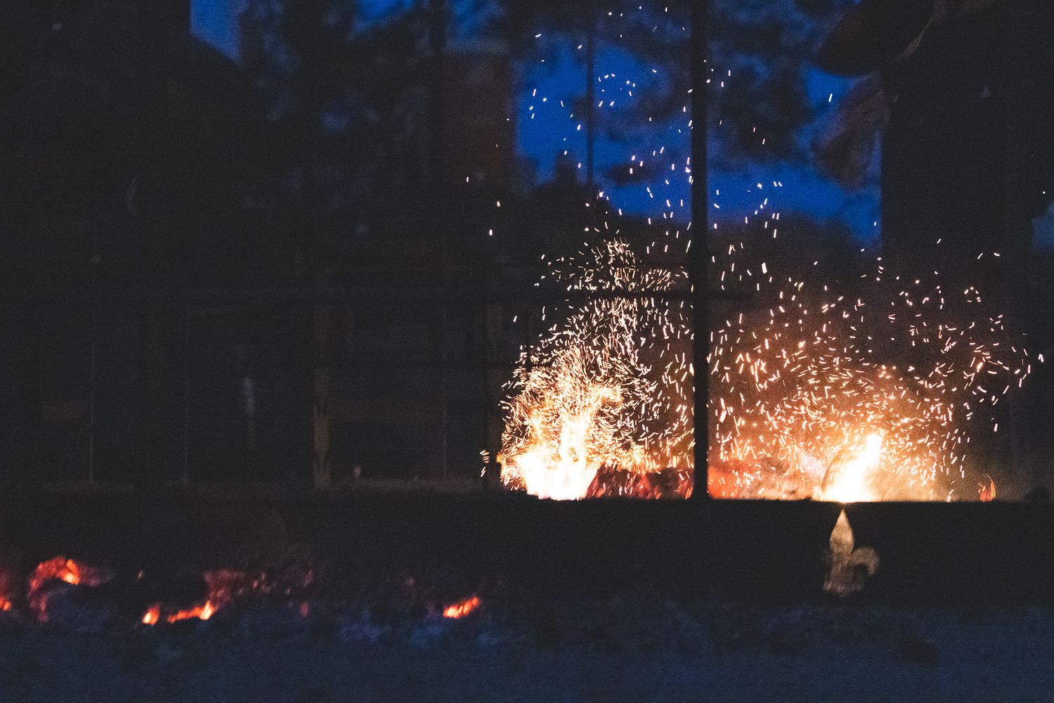 Dramatic nighttime campfire with glowing embers and sparks flying against dark trees and blue sky.