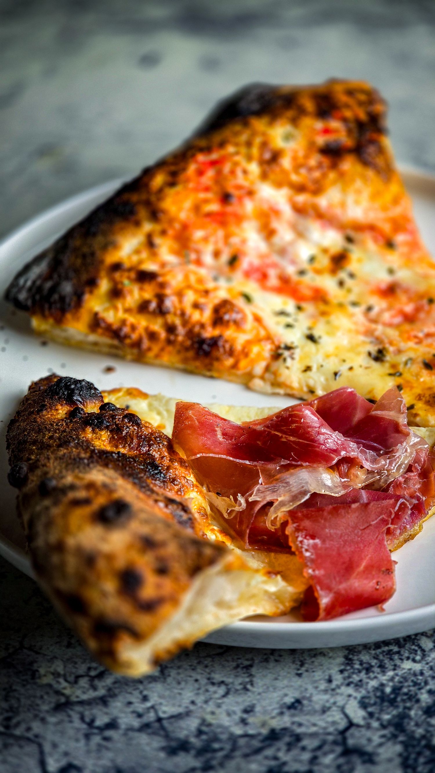 Two slices of Neapolitan pizza with charred crust, melted cheese, tomato sauce, and topped with thin prosciutto on a white plate.