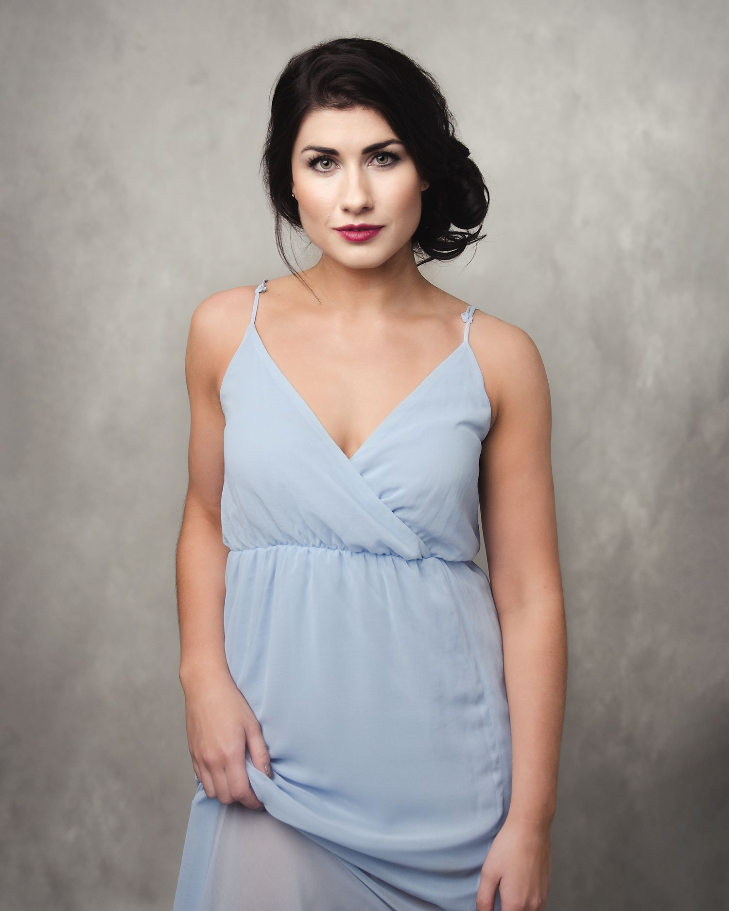 A figure in a light blue sleeveless dress poses against a textured grey backdrop in a studio setting.