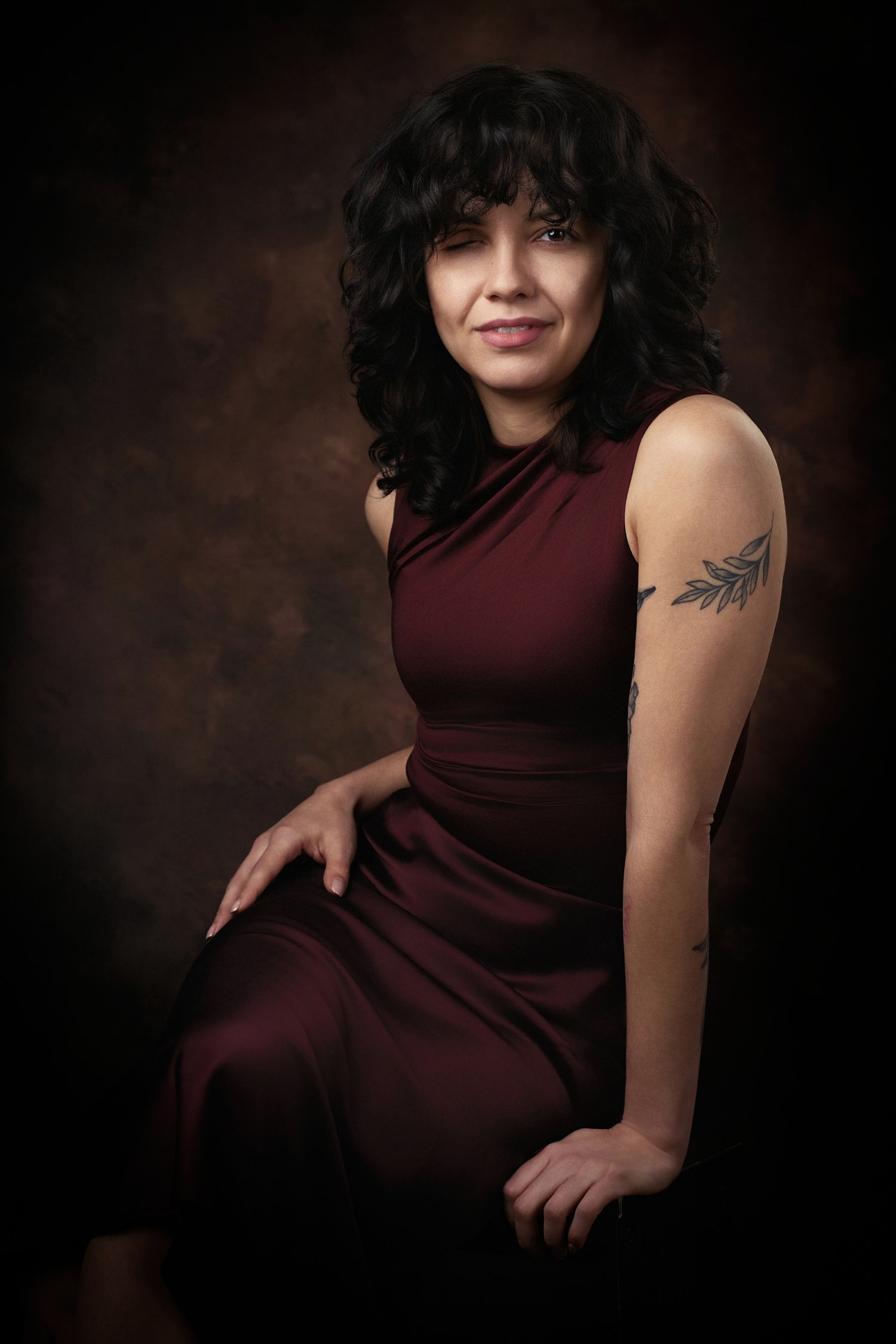 Studio portrait with dark burgundy dress and visible wing tattoo on arm against moody brown background.