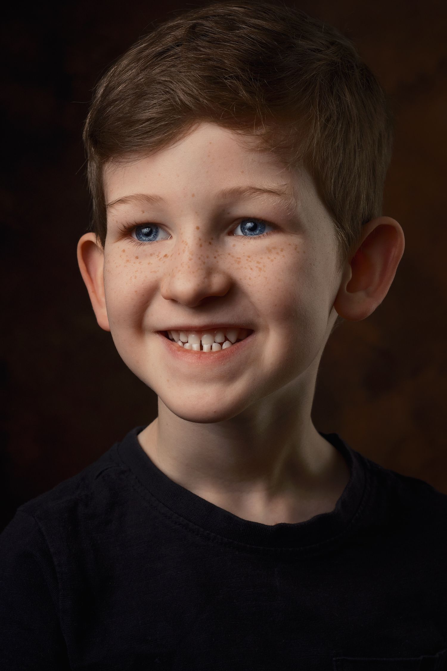 Portrait of a young child with bright blue eyes, freckles, and a cheerful smile wearing a black shirt against a dark background.