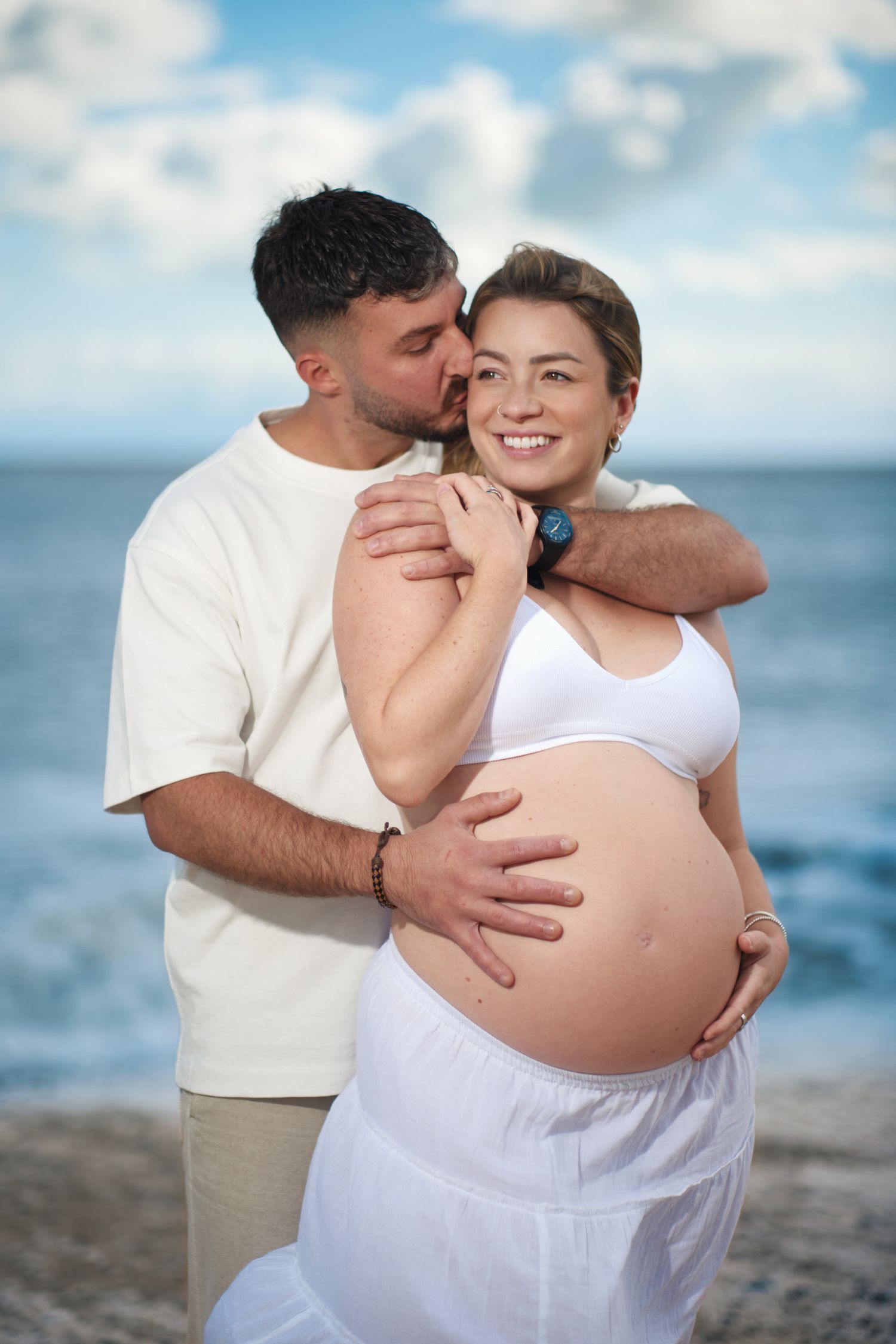 Expectant couple shares intimate moments on a beach during maternity photoshoot with ocean backdrop.