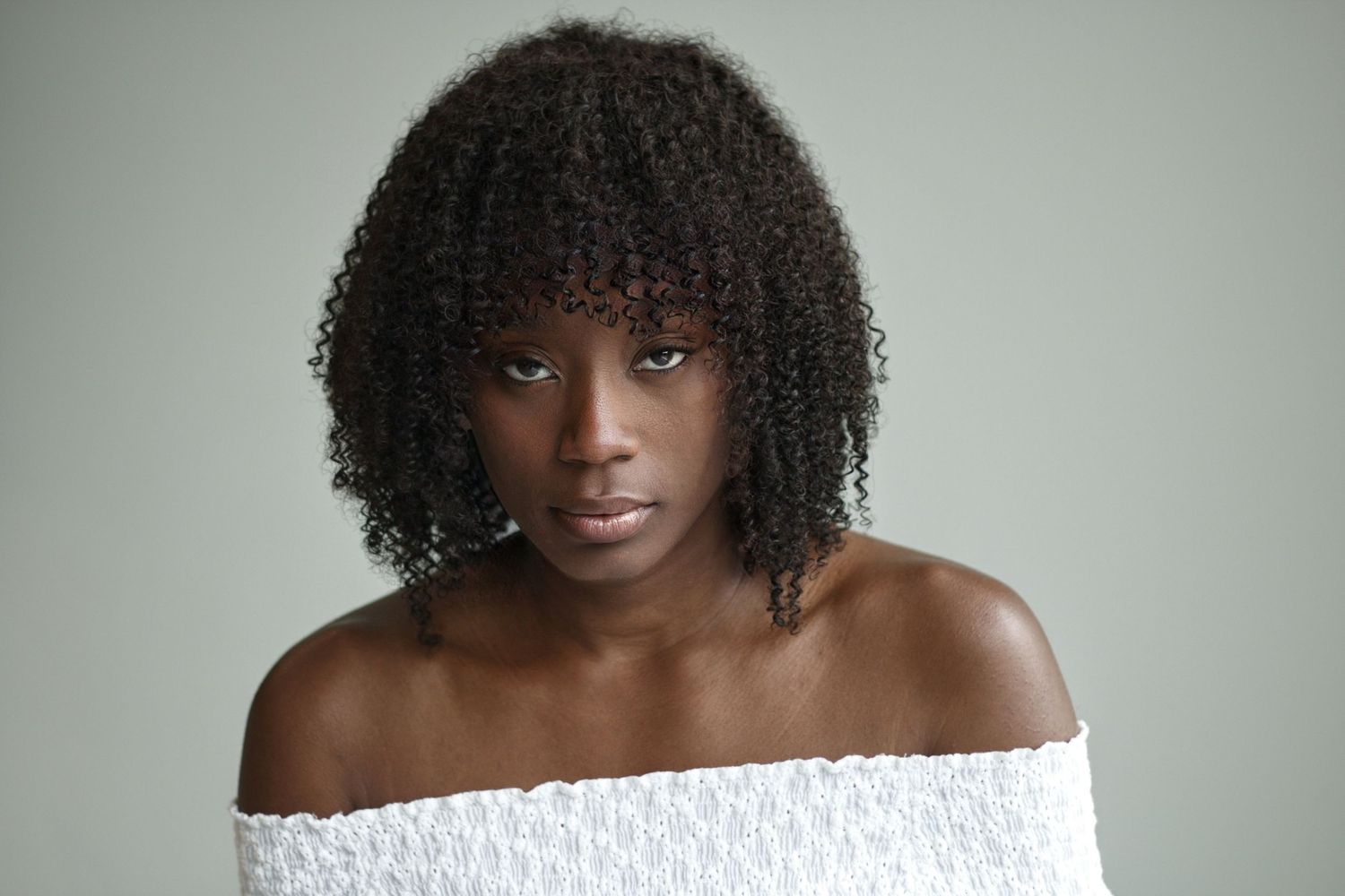 Portrait of a woman with natural curly hair wearing an off-shoulder white top against grey background.