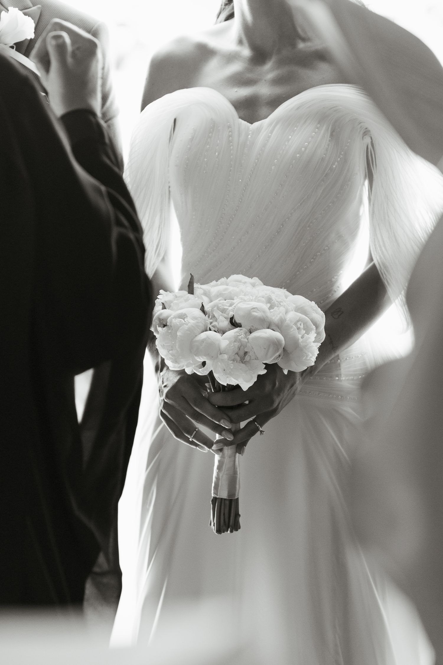 Close-up of white wedding dress and bouquet of white peonies in black and white.