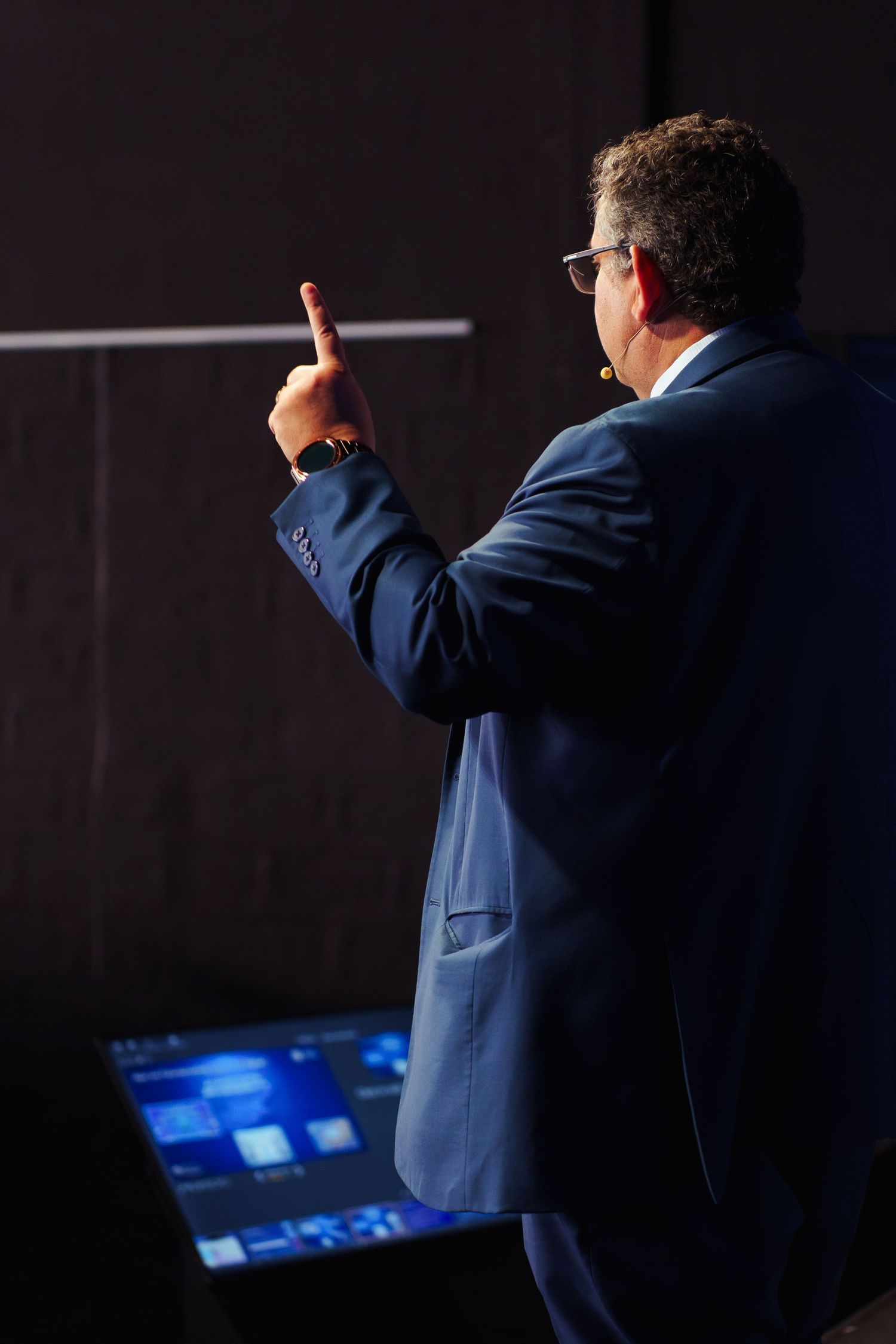 Silhouette of presenter gesturing on dark stage with blue screen in background.