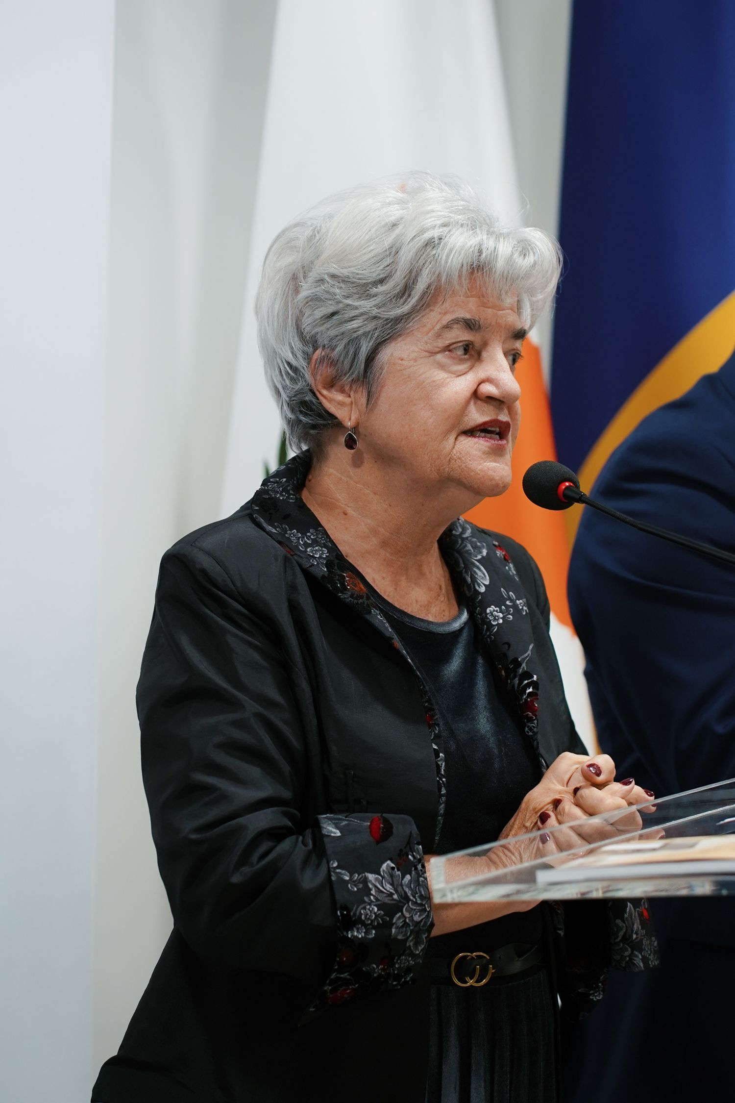 An elderly woman in a black embellished outfit speaks at a podium during a formal event.