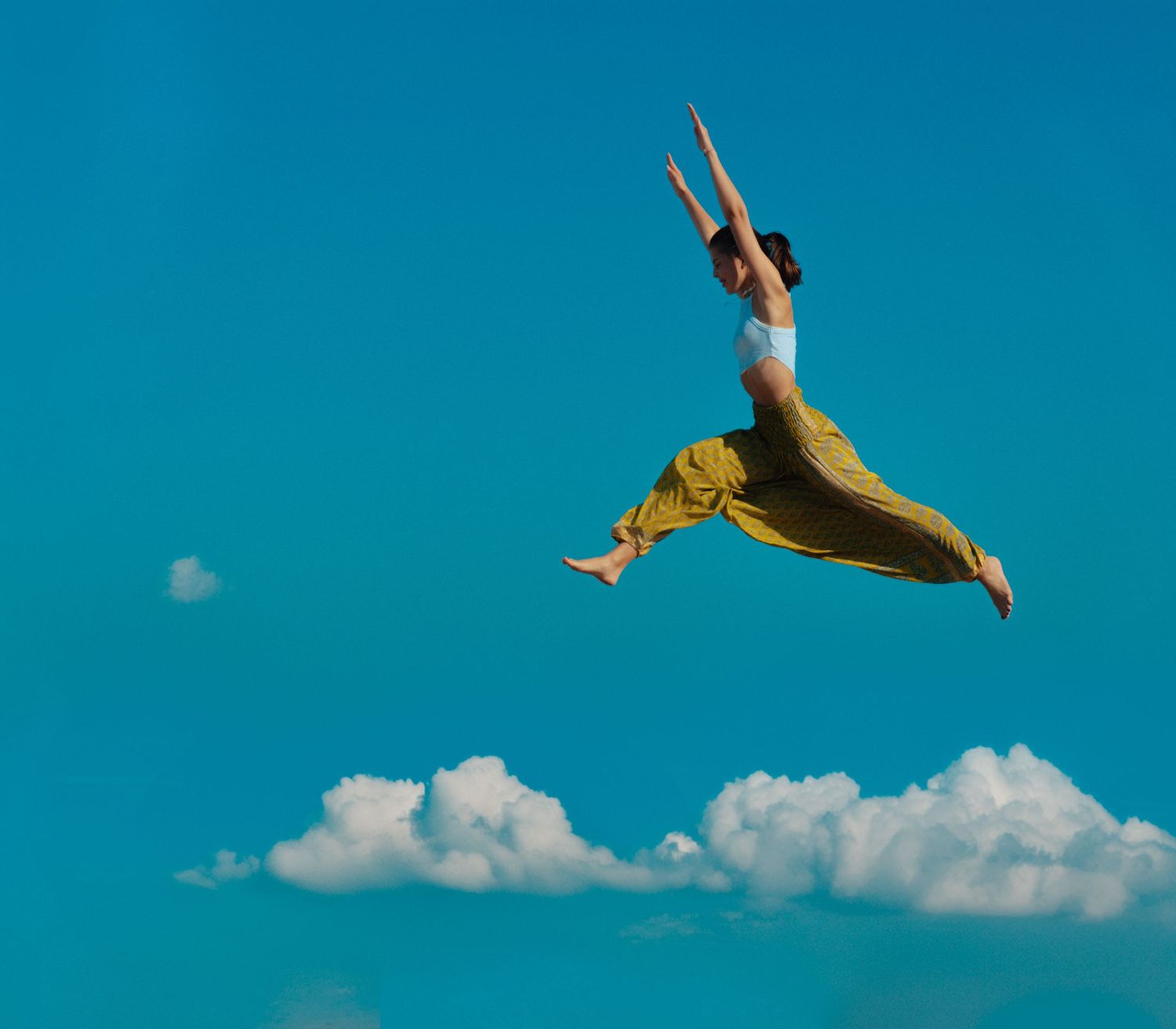 Figure in yellow outfit leaping gracefully against bright blue sky with white clouds.