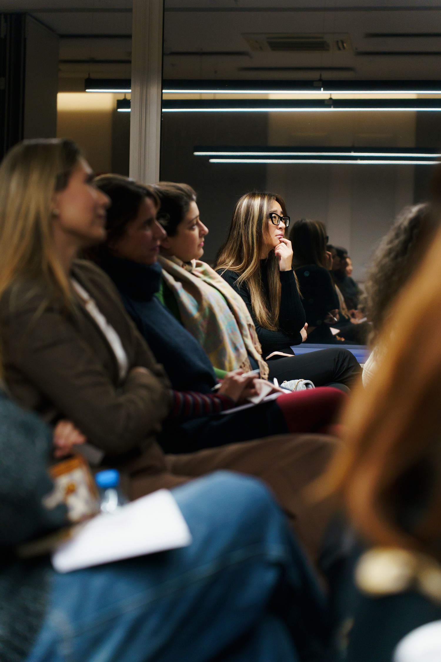 Students sit attentively in rows during a lecture or presentation in a dimly lit classroom setting.