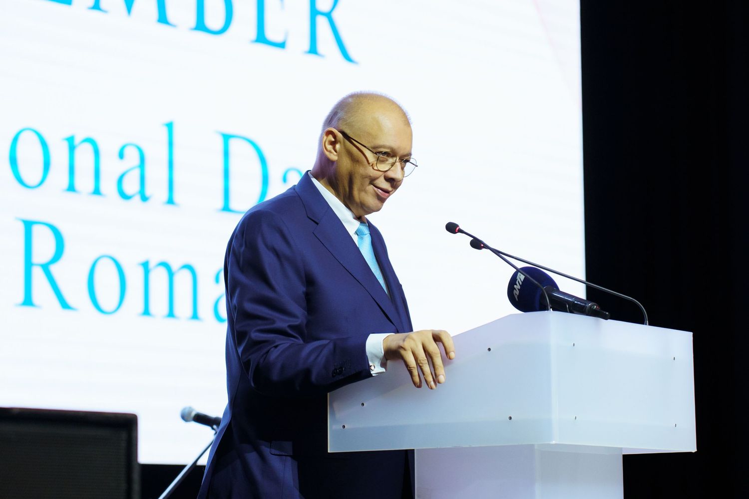 Speaker presenting at a podium during a formal event with text display in background.