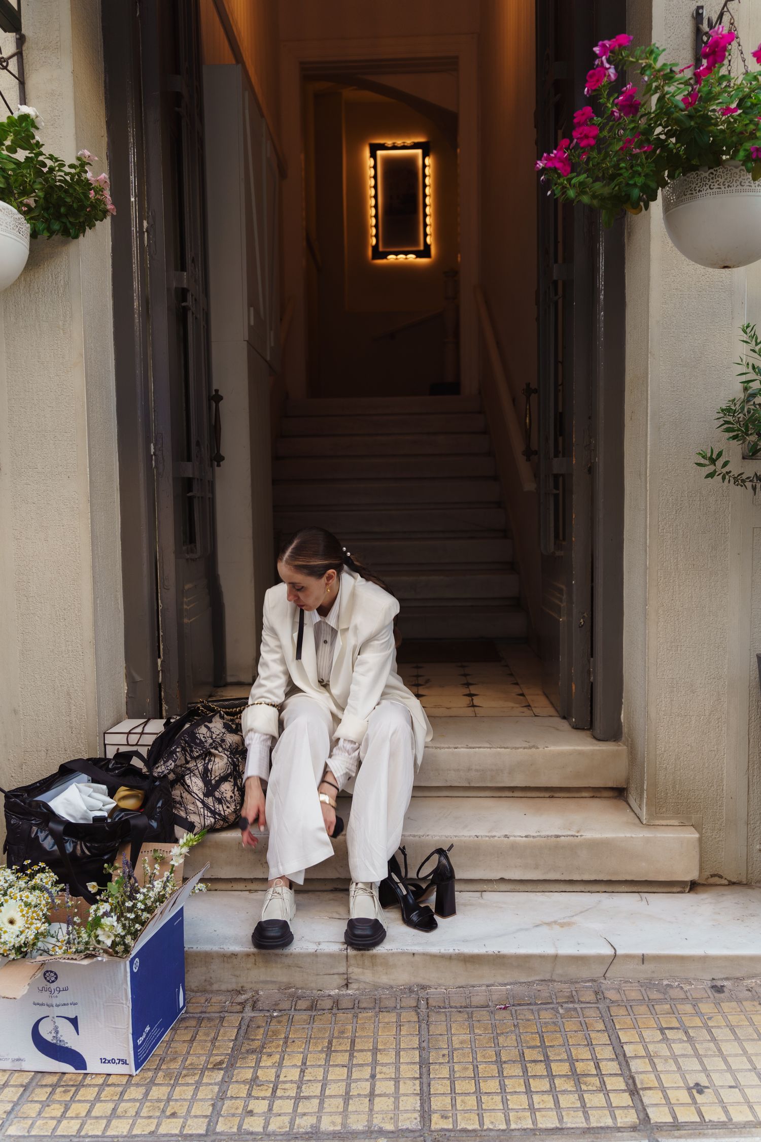 Person in white suit sitting alone on steps with luggage and shoes beside them.