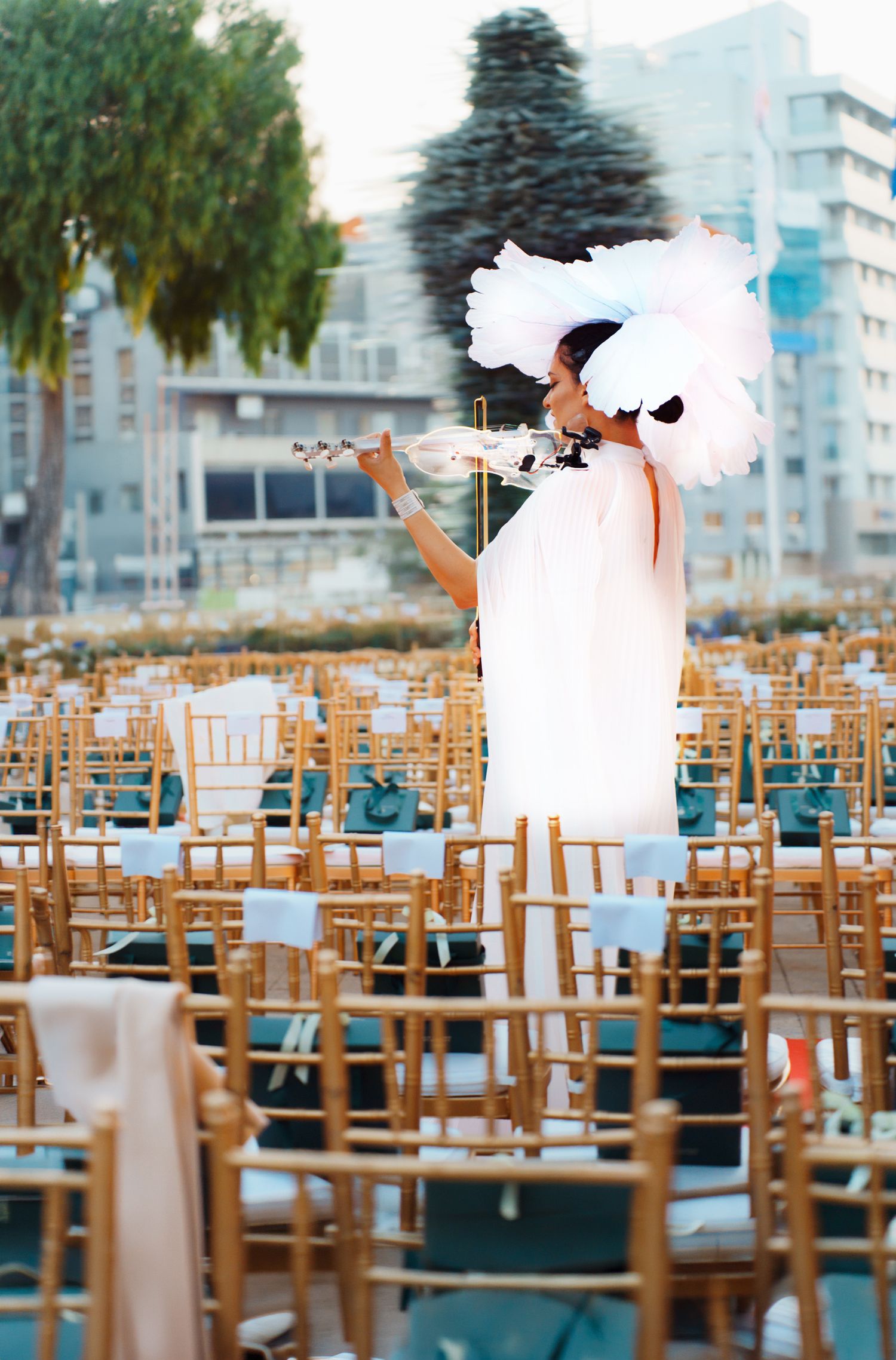 Violinist performs in white dress among empty chairs at an outdoor venue setup.