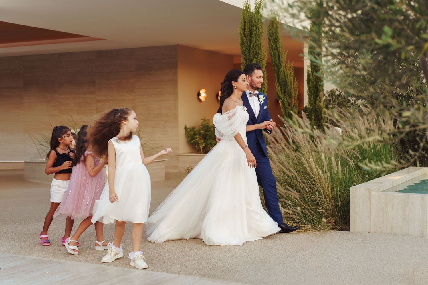 Wedding party members in formal attire walking along an outdoor pathway near modern architecture.