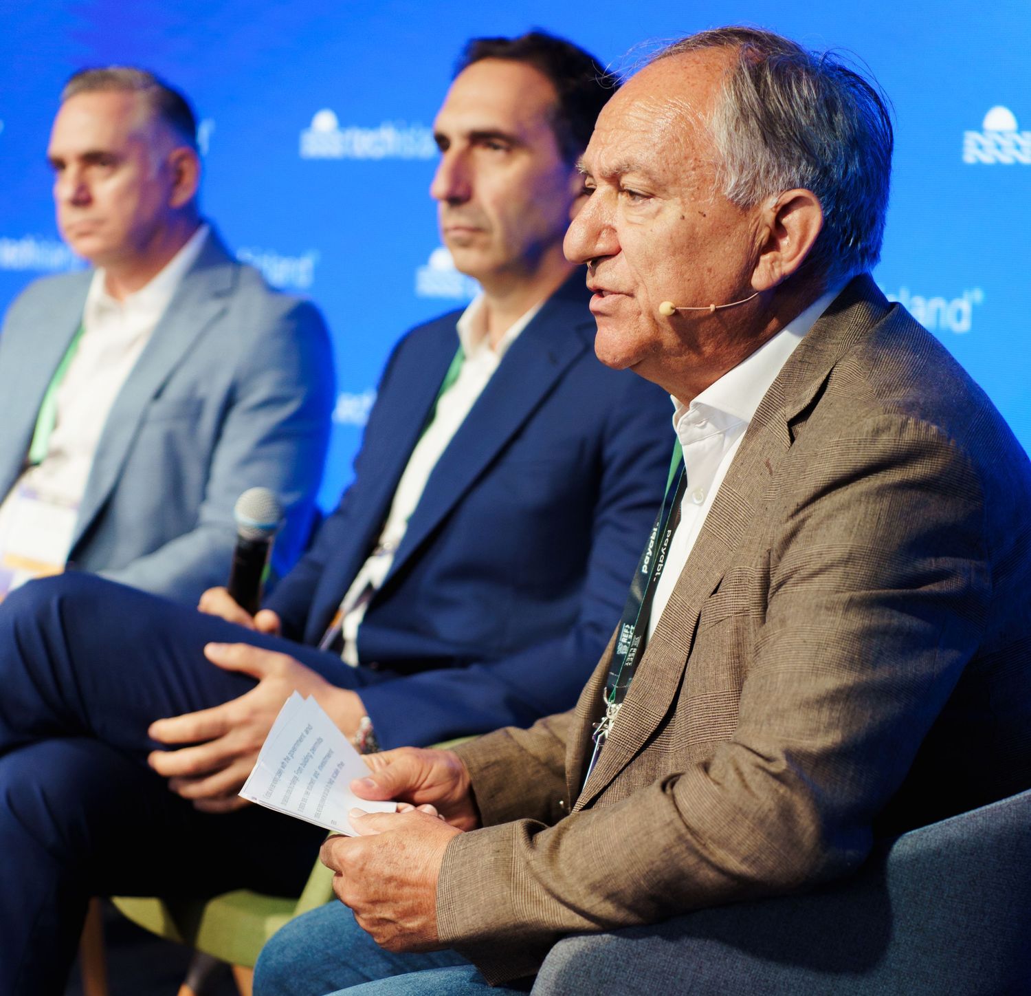 Three men in business attire seated on stage during a panel discussion at a professional event.